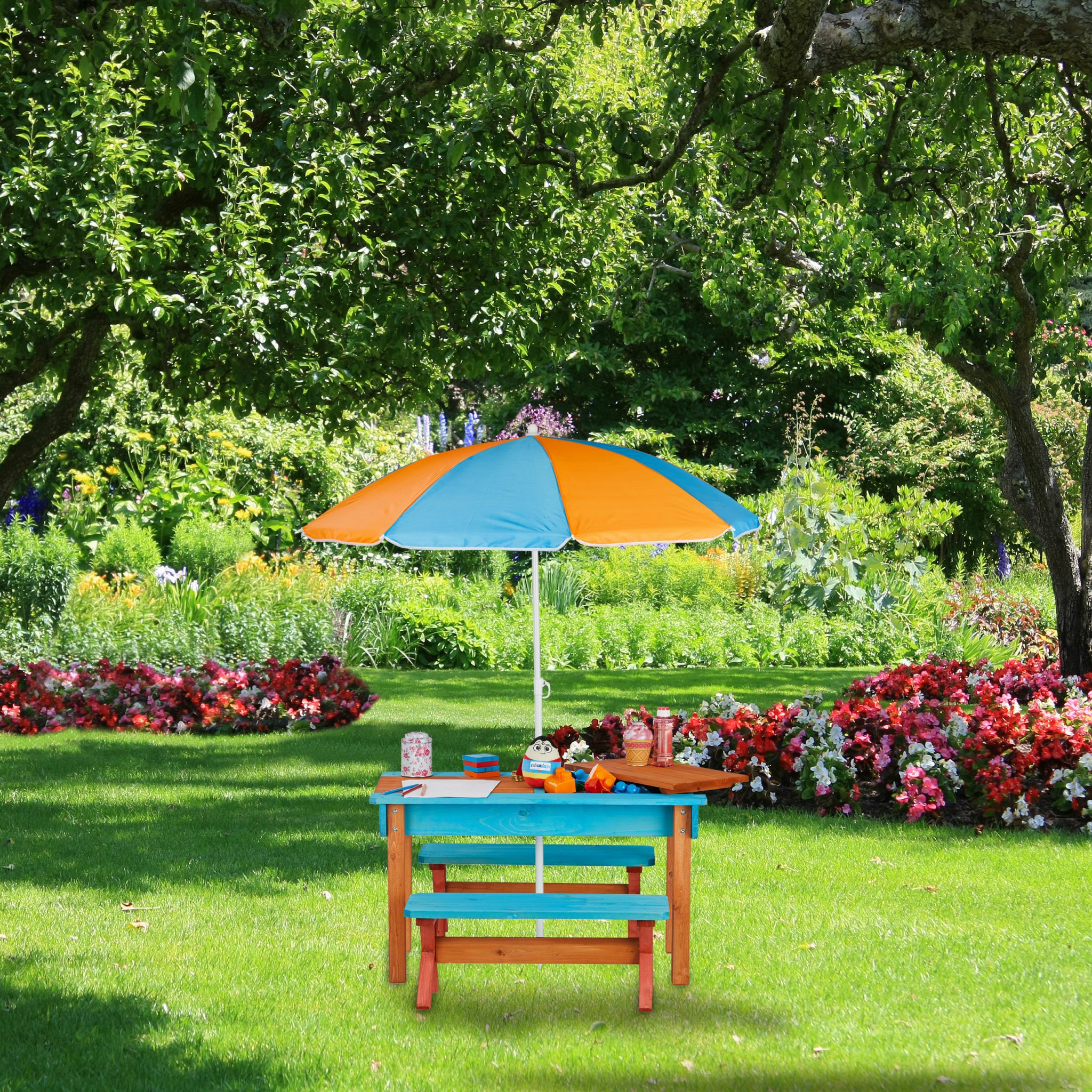 Ein blauer Kindertisch mit Regenschirm steht auf einem grünen Rasen in einem blühenden Garten.