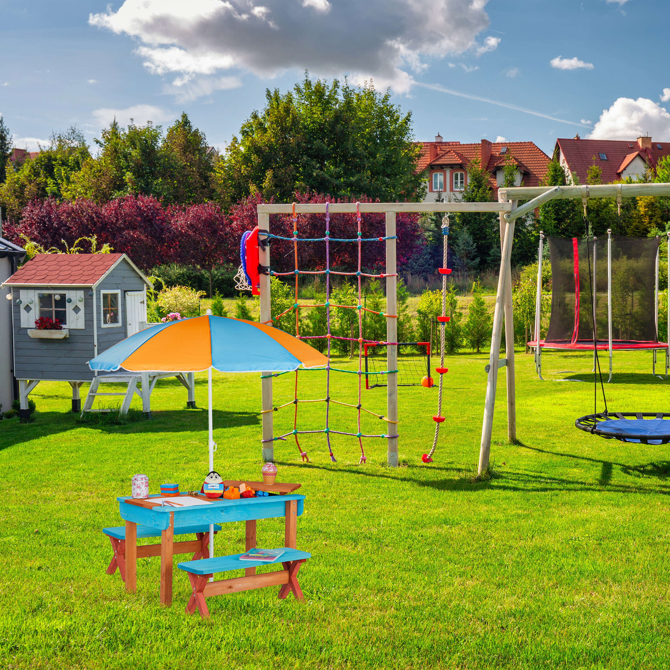 Bunter Garten mit Spielhaus, Klettergerüst, Trampolin und einem blauen Kindertisch mit Sonnenschirm.