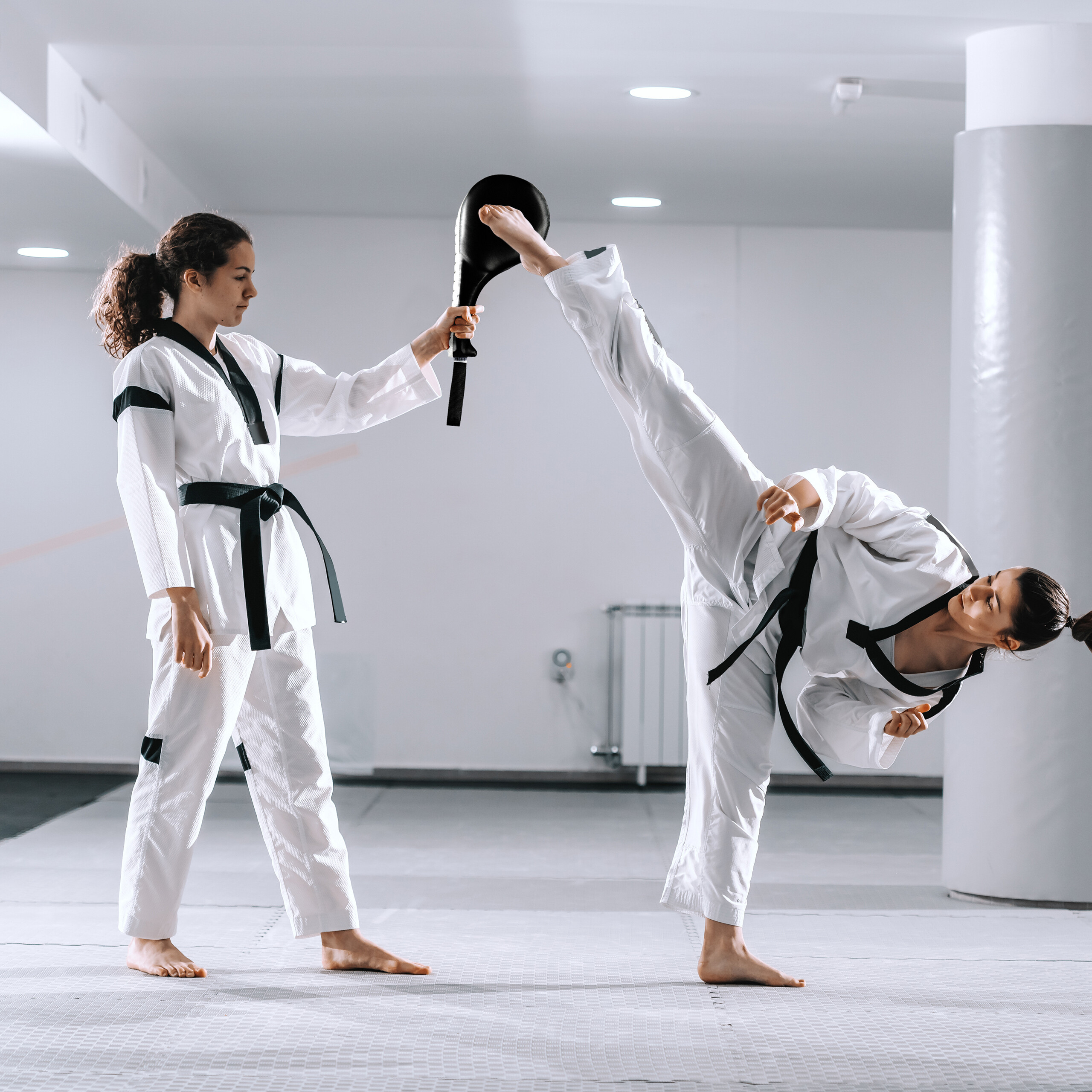 Zwei Frauen in Taekwondo-Uniformen üben in einer hellen Halle einen Tritt gegen ein Ziel.