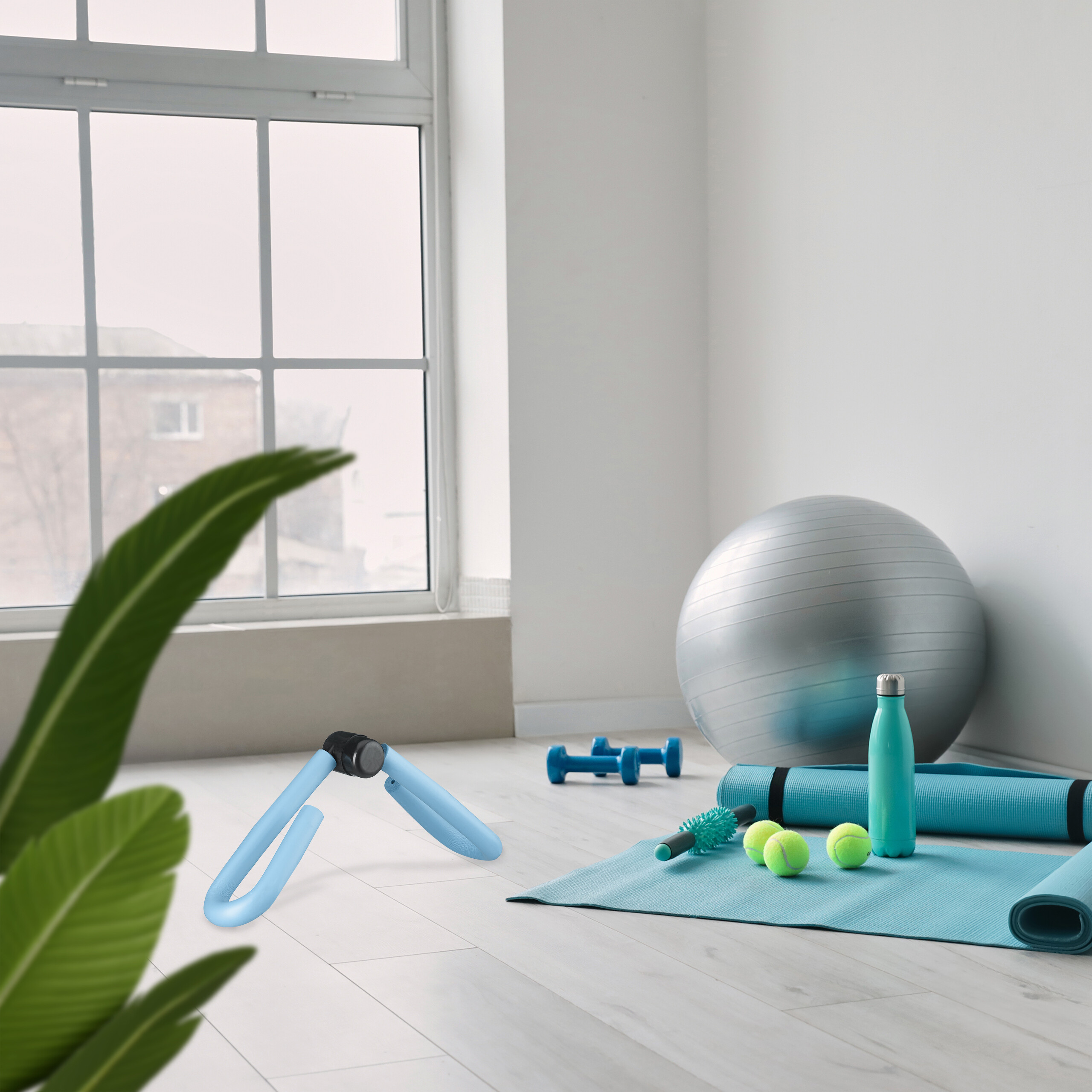 Fitnessgeräte in hellem Raum mit Fenster, Übungsball, blaue Matte, Gewichte und grüne Tennisbälle auf dem Boden.