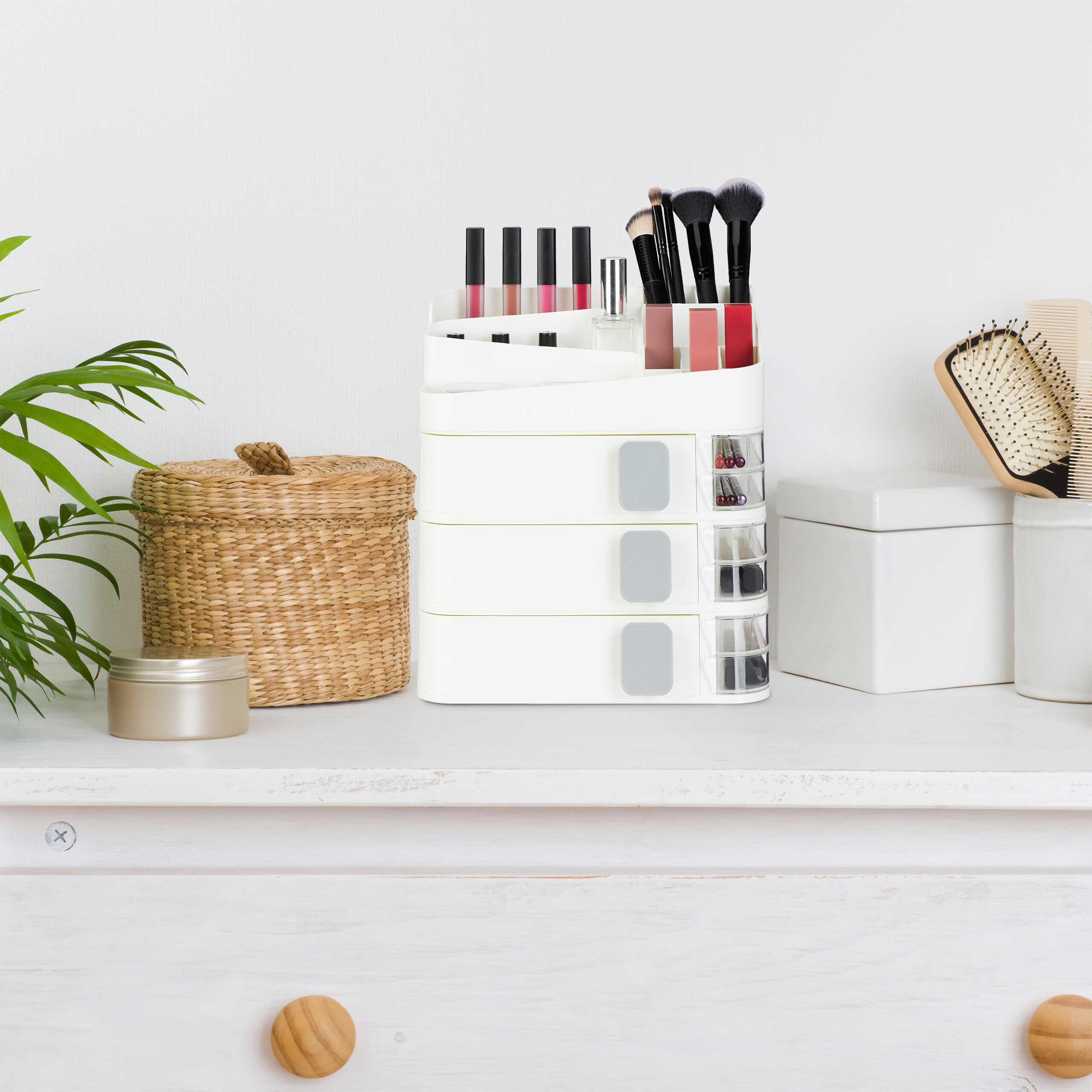 Eine weiße Make-up-Schublade mit Pinseln und Lippenstiften auf einer weißen Kommode neben einem Korb.