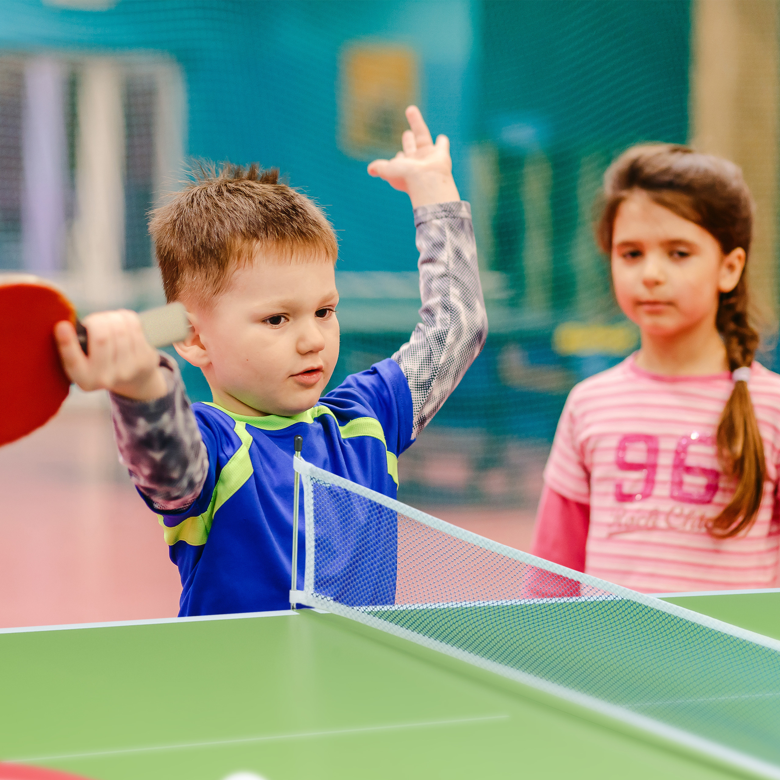 Ein kleiner Junge im blauen T-Shirt spielt Tischtennis, während ein Mädchen im rosa zuschaut.