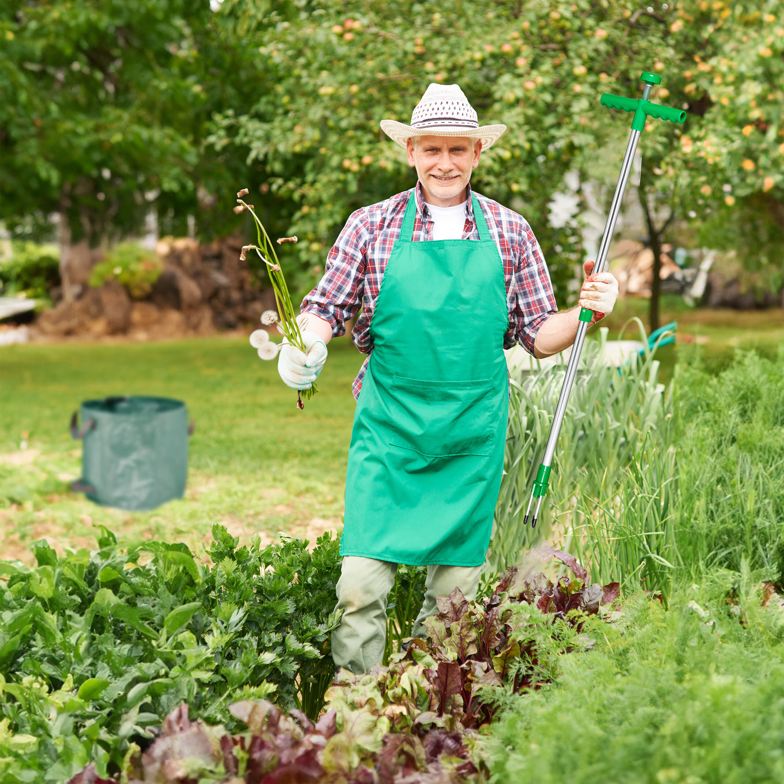 Lächelnder Gärtner im Hut und gr üner Schürze hält Unkraut und ein Unkrautentfernungsgerät im Garten.
