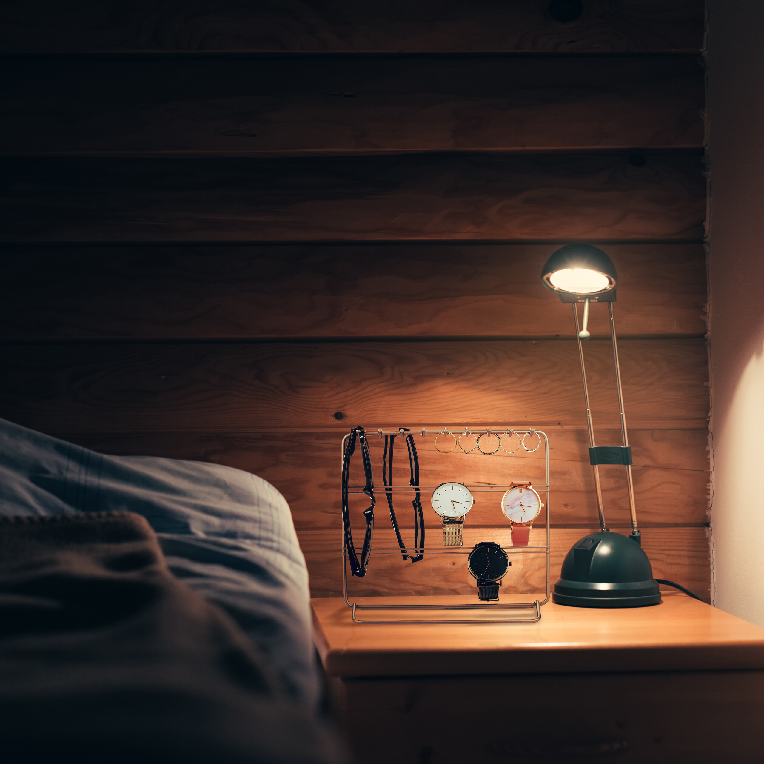 Eine Nachttischlampe beleuchtet Uhren und Schmuck auf einem Ständer vor einer Holzverkleidung.