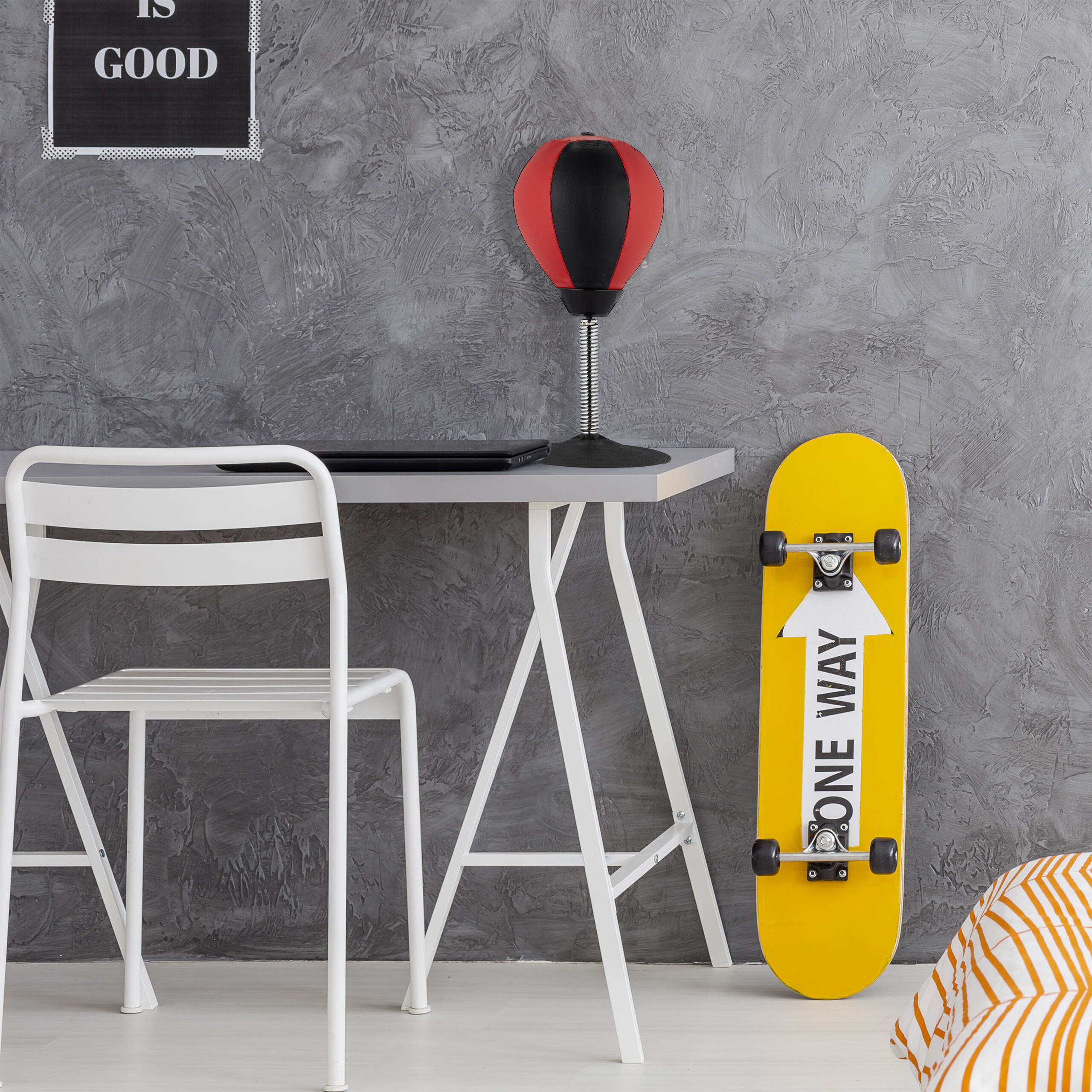 Ein Schreibtisch mit Laptop und Punchingball, daneben ein weißer Stuhl und ein gelbes Skateboard vor einer grauen Wand.