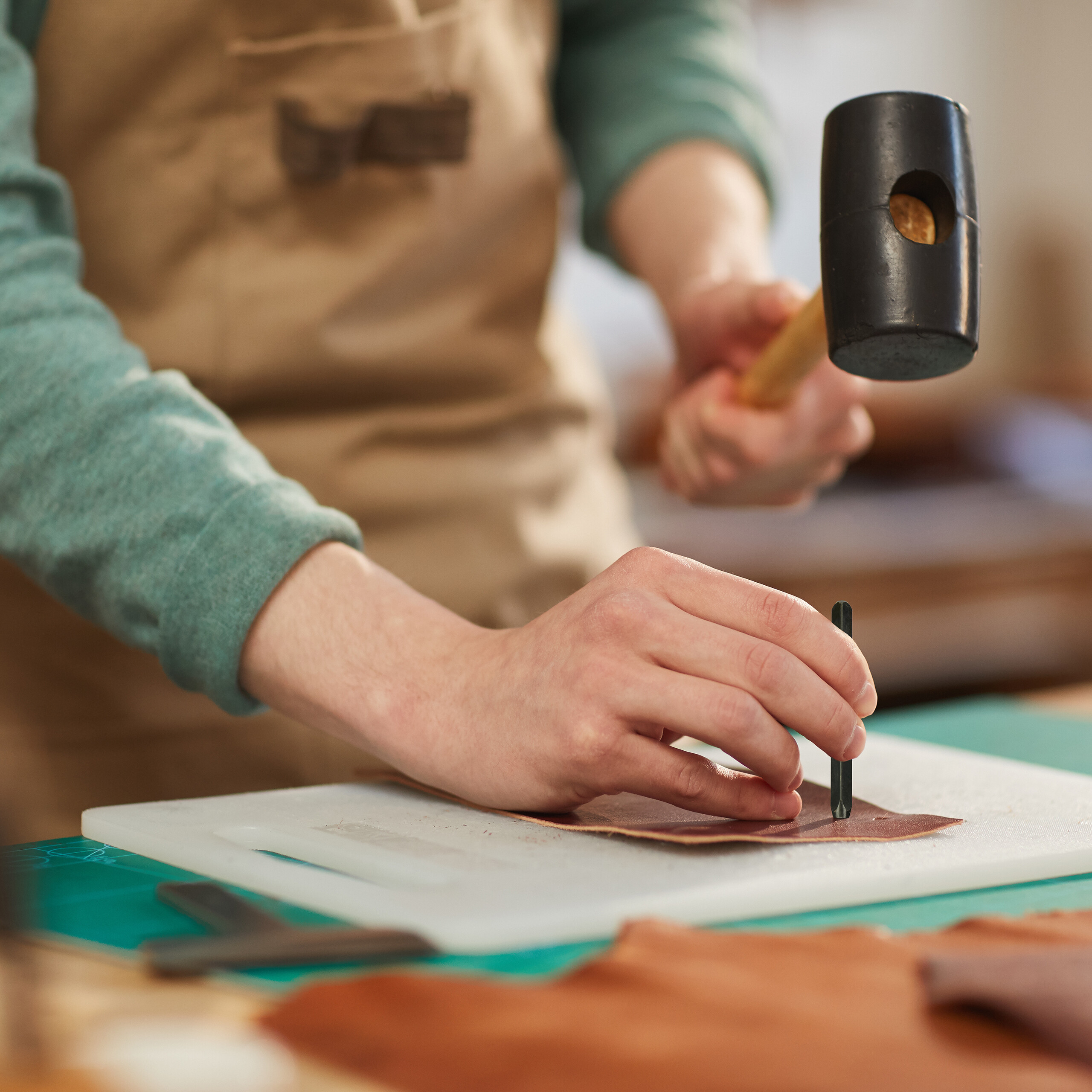 Handwerker mit brauner Schürze schlägt mit Hammer auf Lederstück auf weißem Brett.