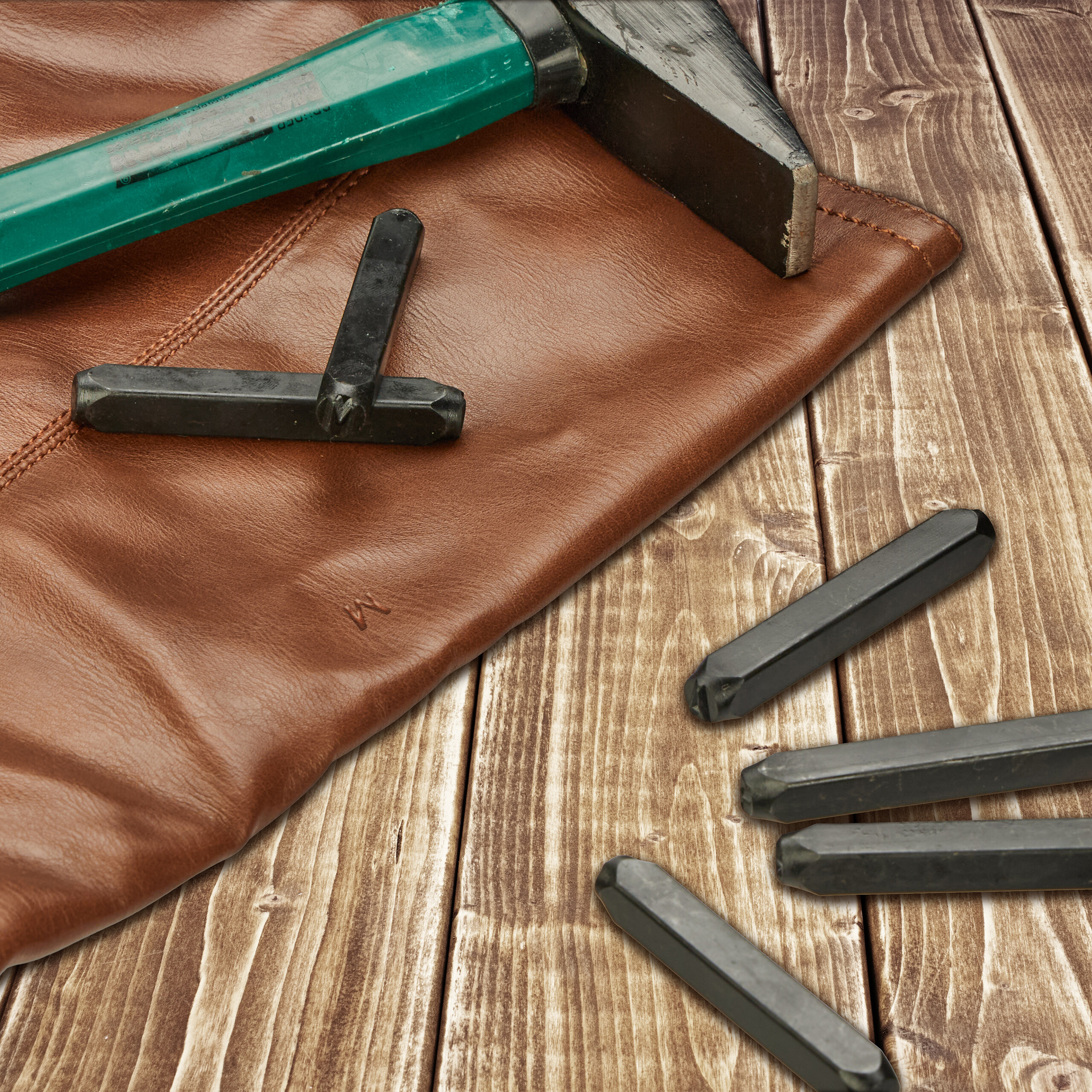 Ein grüner Hammer und schwarze Metallstempel liegen auf braunem Leder und einer Holzoberfläche.