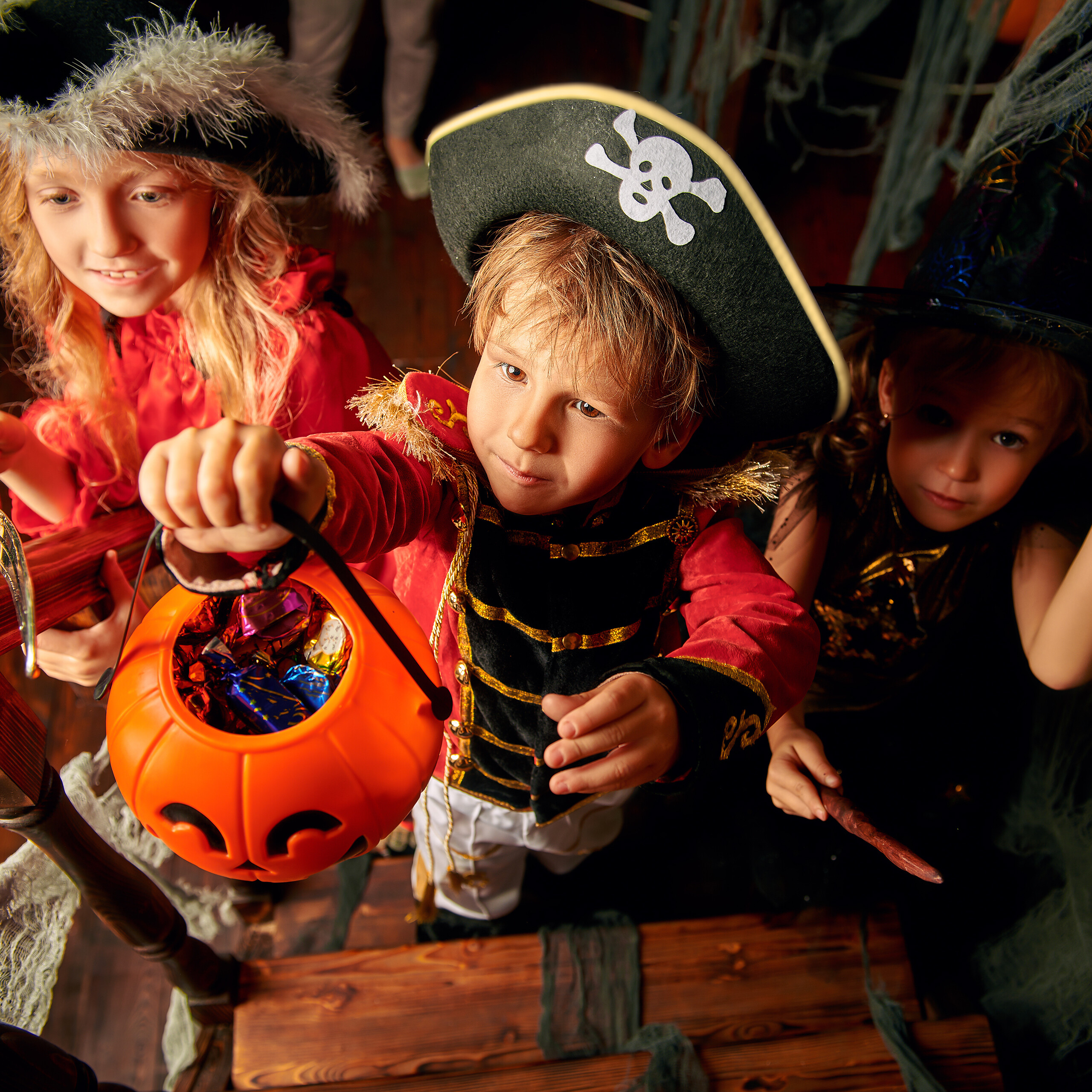Drei Kinder in Halloween-Kostümen halten einen Kürbiseimer voller Süßigkeiten auf einer Treppe.