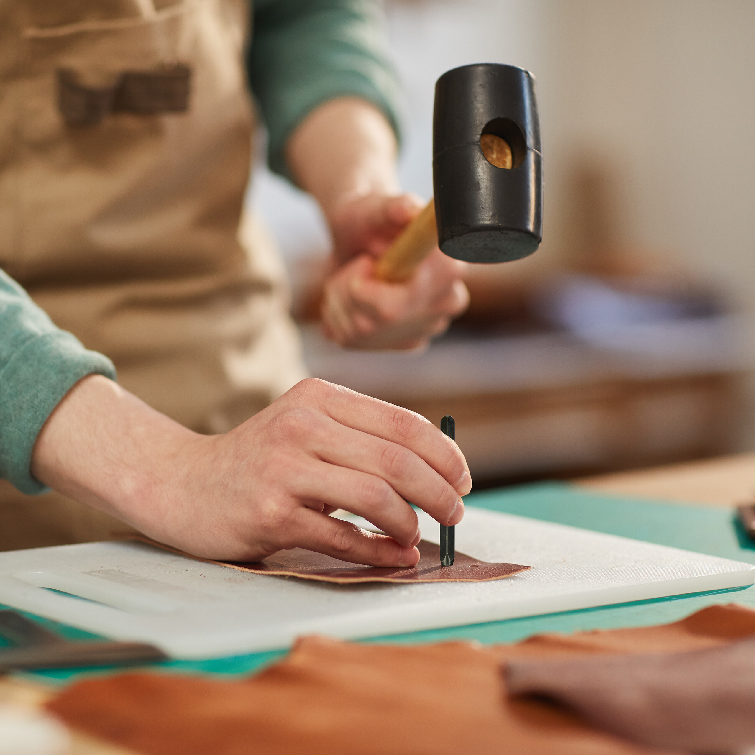 Handwerker mit brauner Schürze schlägt mit einem Gummihammer auf ein Werkzeug, um Leder zu lochen.