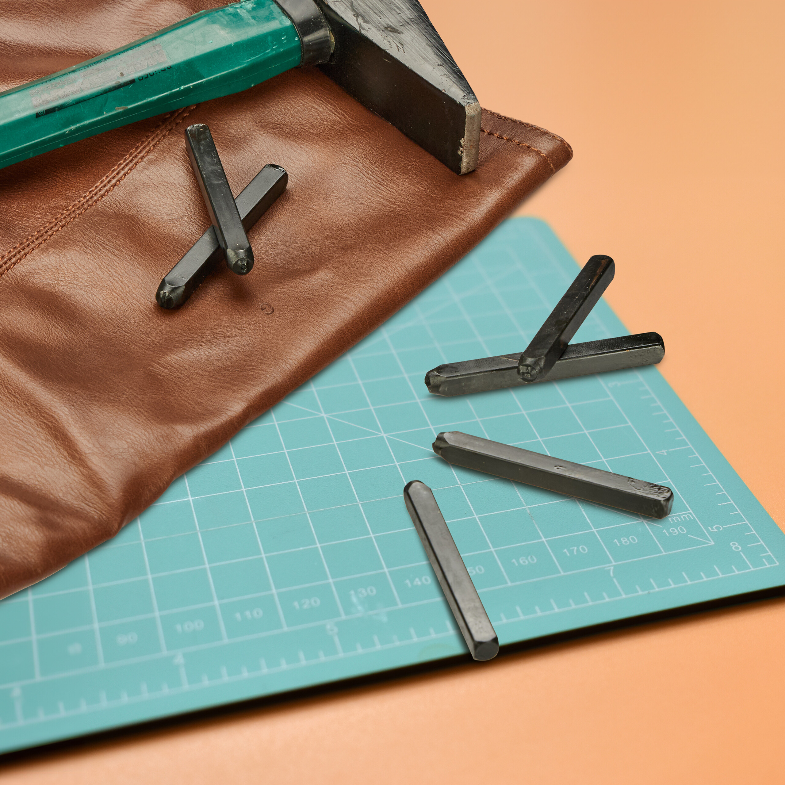 Ein Hammer, braunes Leder und Metallstempel liegen auf einer grünen Schneidematte und orangefarbenem Untergrund.
