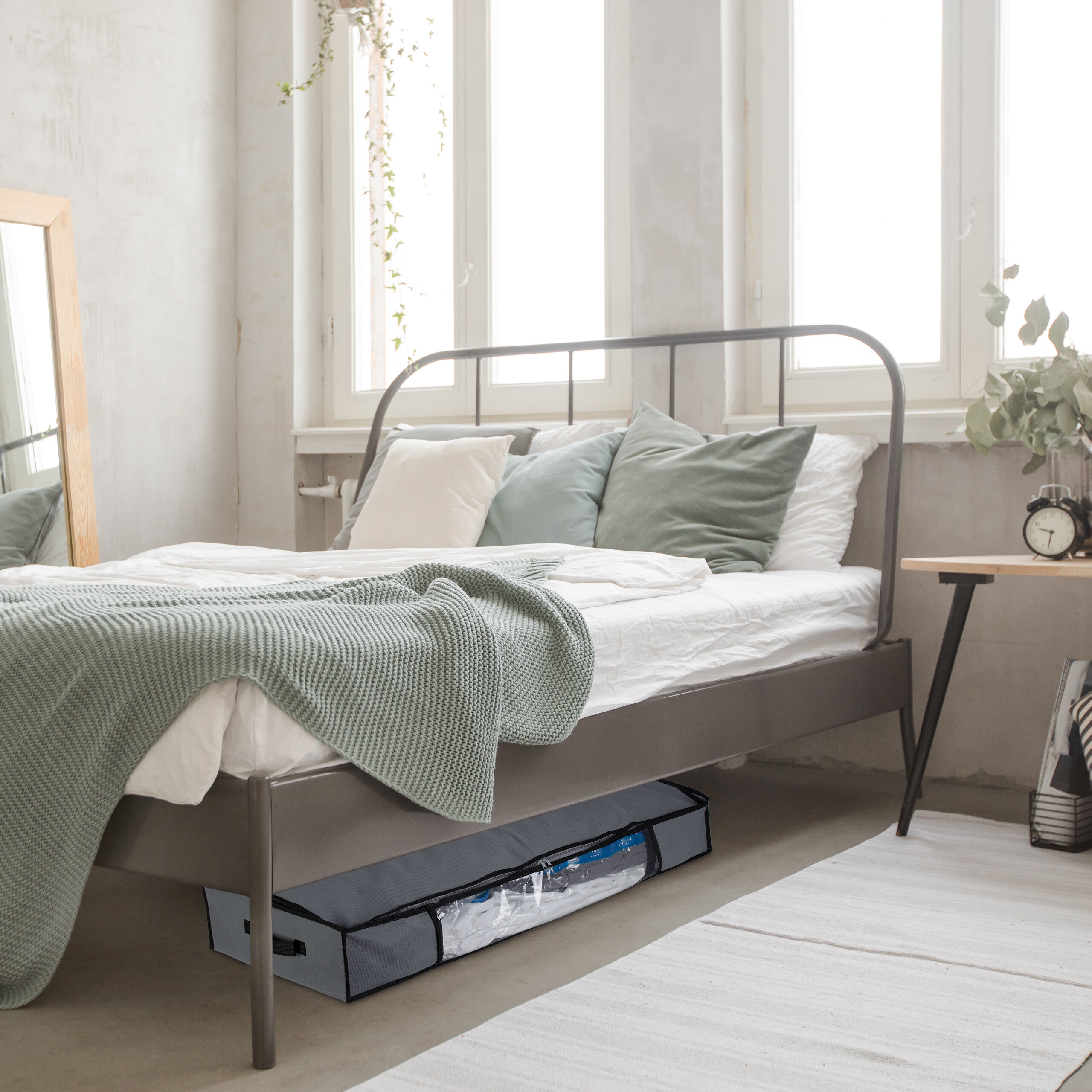 Ein grauer Unterbettkommoden-Aufbewahrungsbeutel lagert unter dem Bettgestell in einem hellen Schlafzimmer.