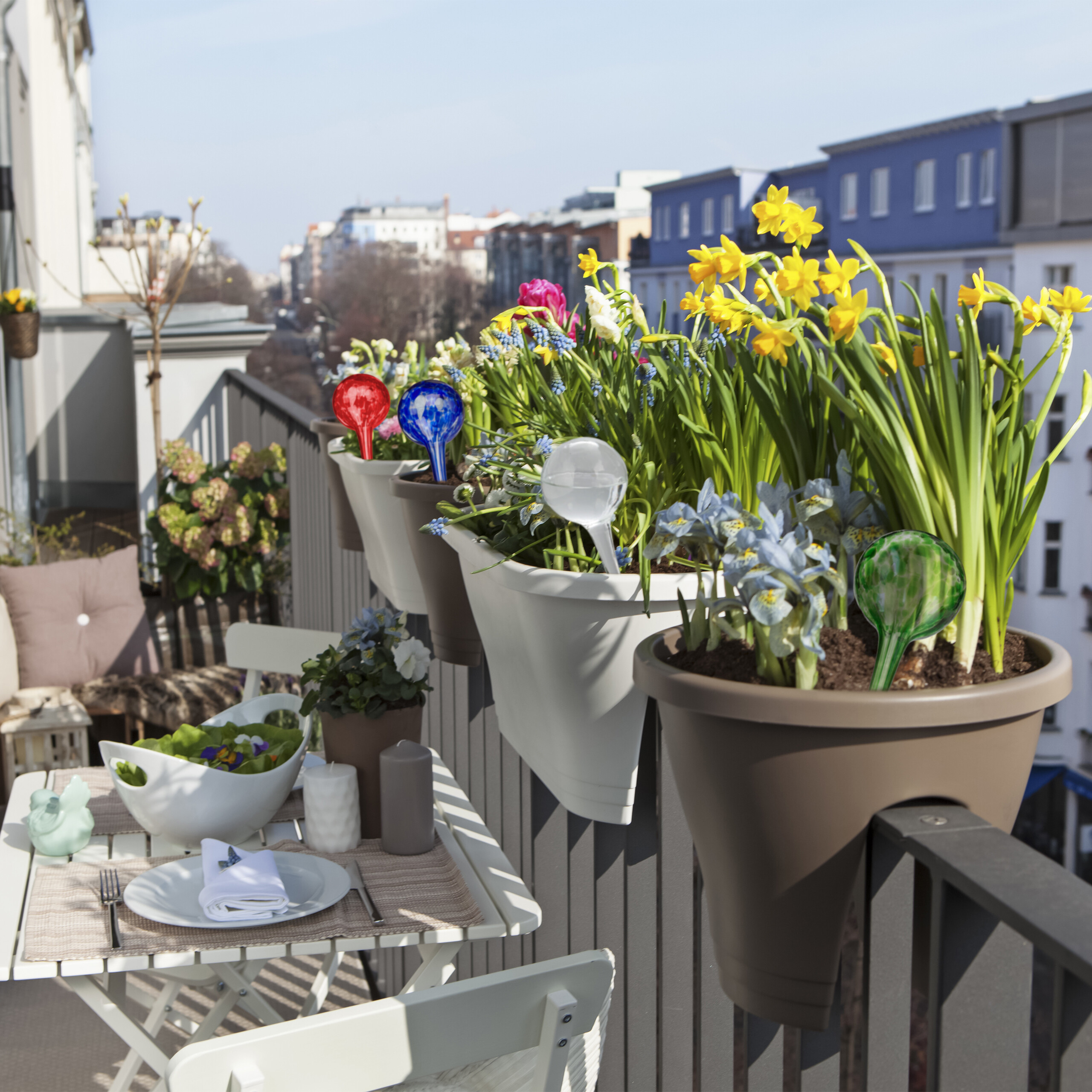 Ein sonniger Balkon mit Blumenkästen voller gelber Narzissen, bunten Glas-Wasserspendern und einem gedeckten Tisch.