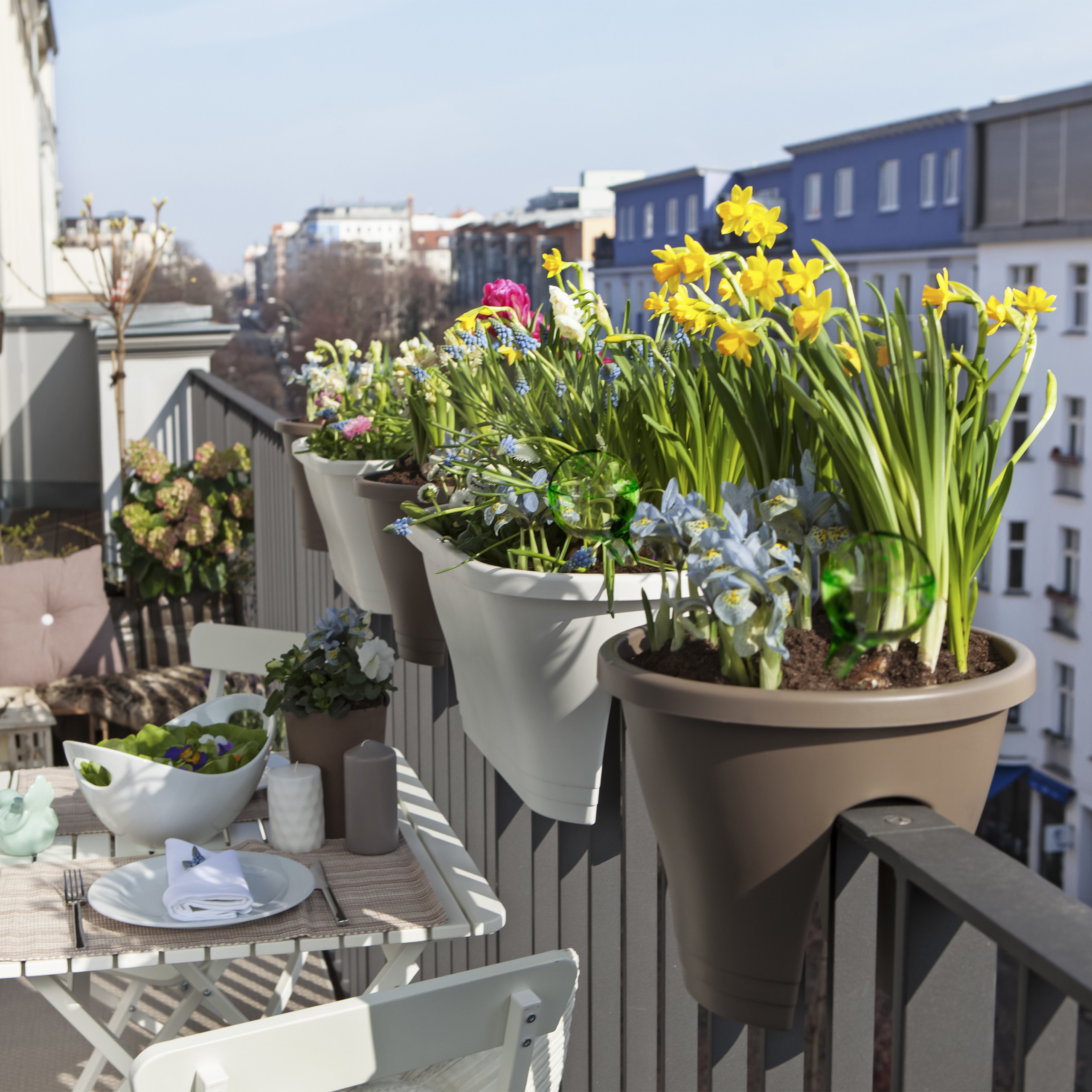 Ein sonniger Balkon mit Blumentöpfen am Geländer und einem gedeckten Tisch mit Blick auf die Stadt.