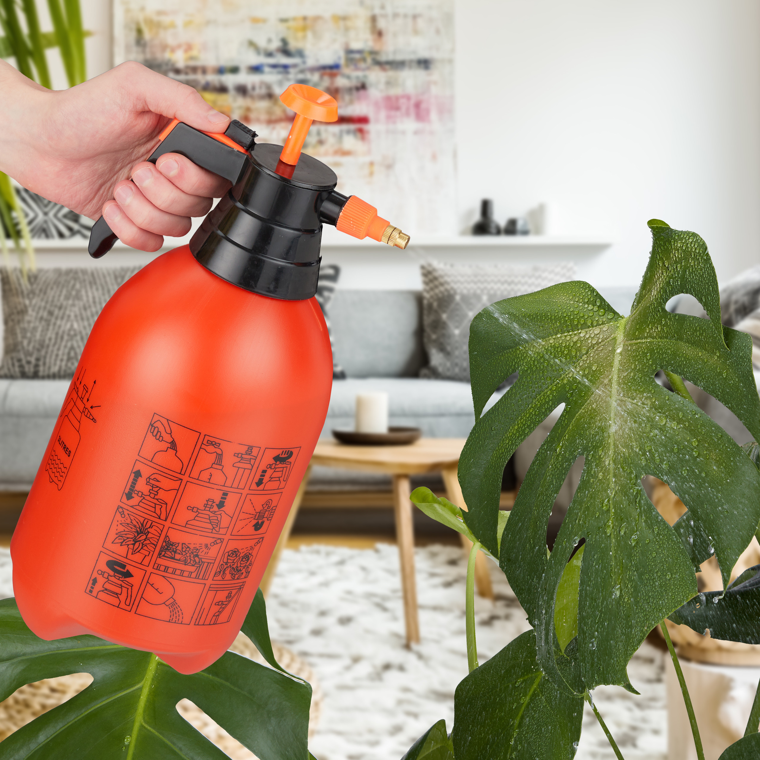 Eine Hand hält einen orangefarbenen Pumpsprüher und besprüht eine nasse Monstera-Pflanze in einem hellen Wohnzimmer.