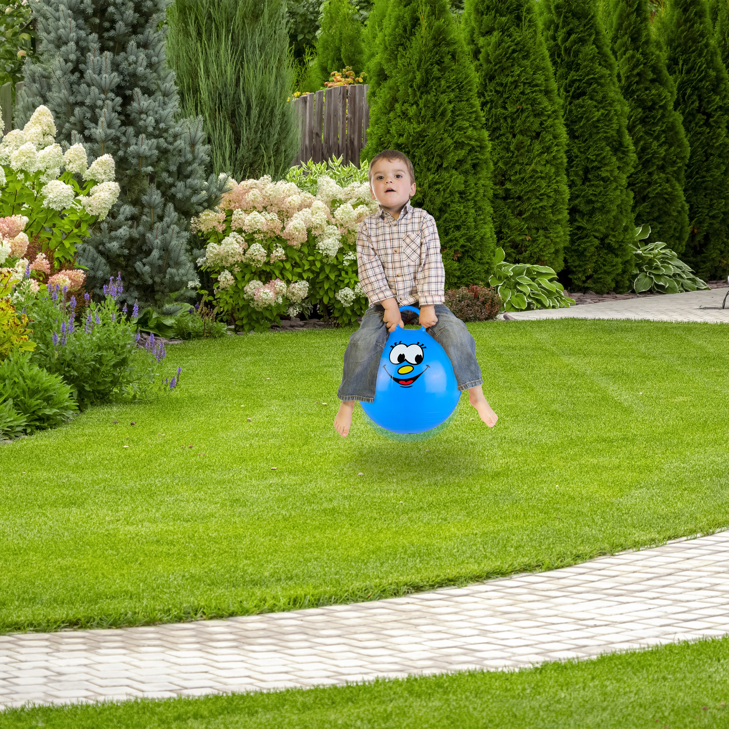 Ein Junge hüpft glücklich auf einem blauen Hüpfball mit Smiley in einem üppig grünen Garten.