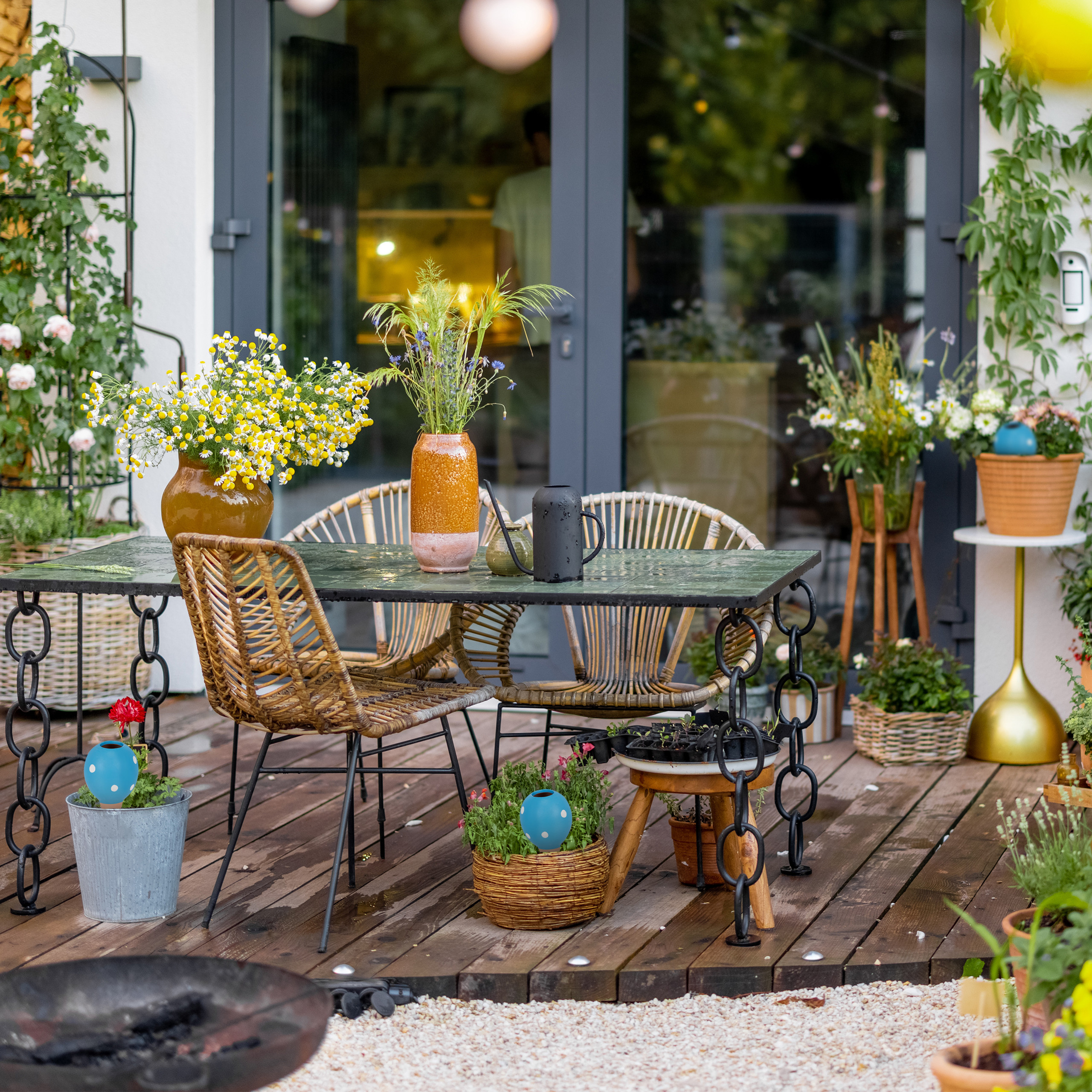 Holzterrasse mit Esstisch, Rattanstühlen und vielen bunten Blumen in Vasen und Töpfen vor einem Haus.