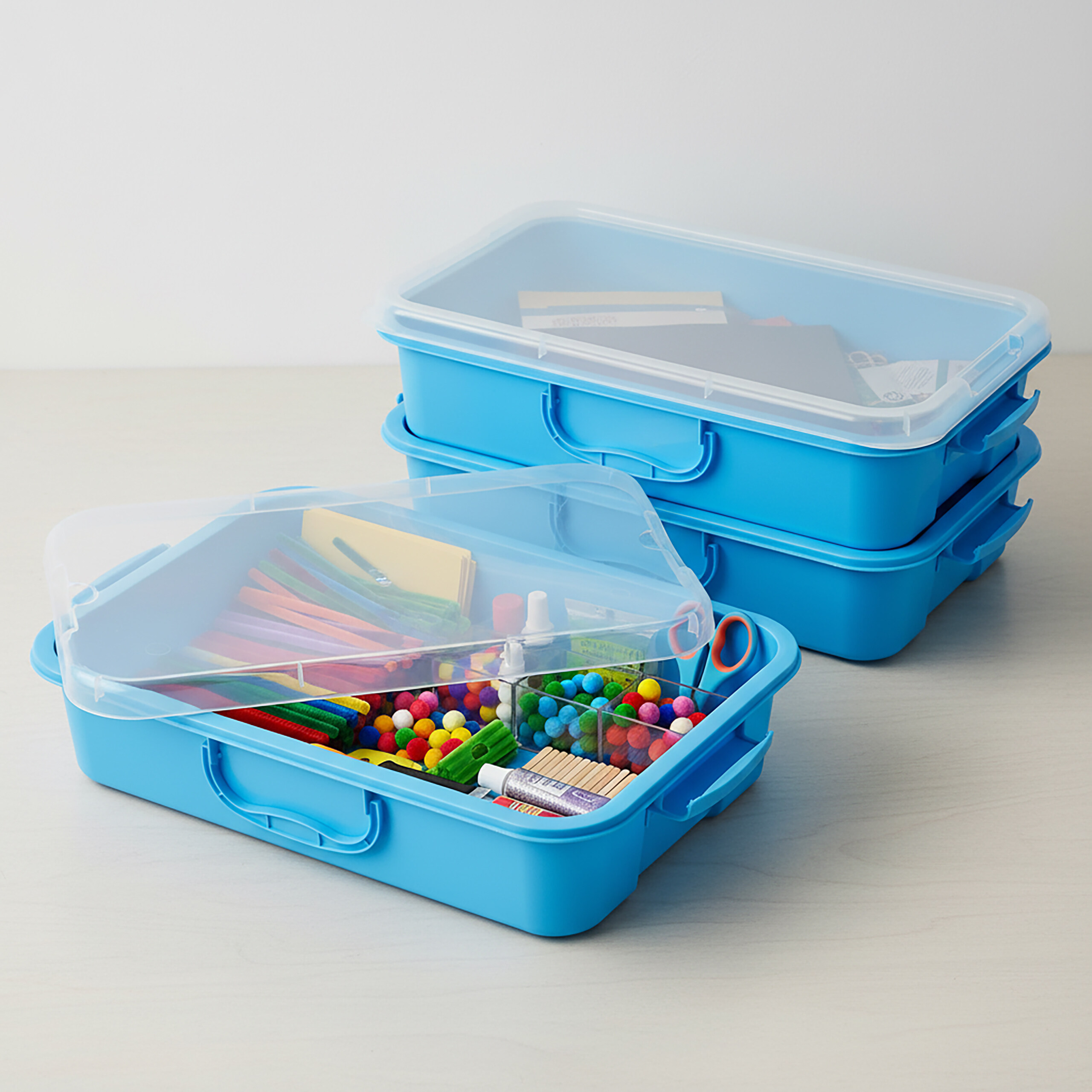 Drei blaue Kunststoffboxen mit transparenten Deckeln, gefüllt mit bunten Bastelmaterialien.