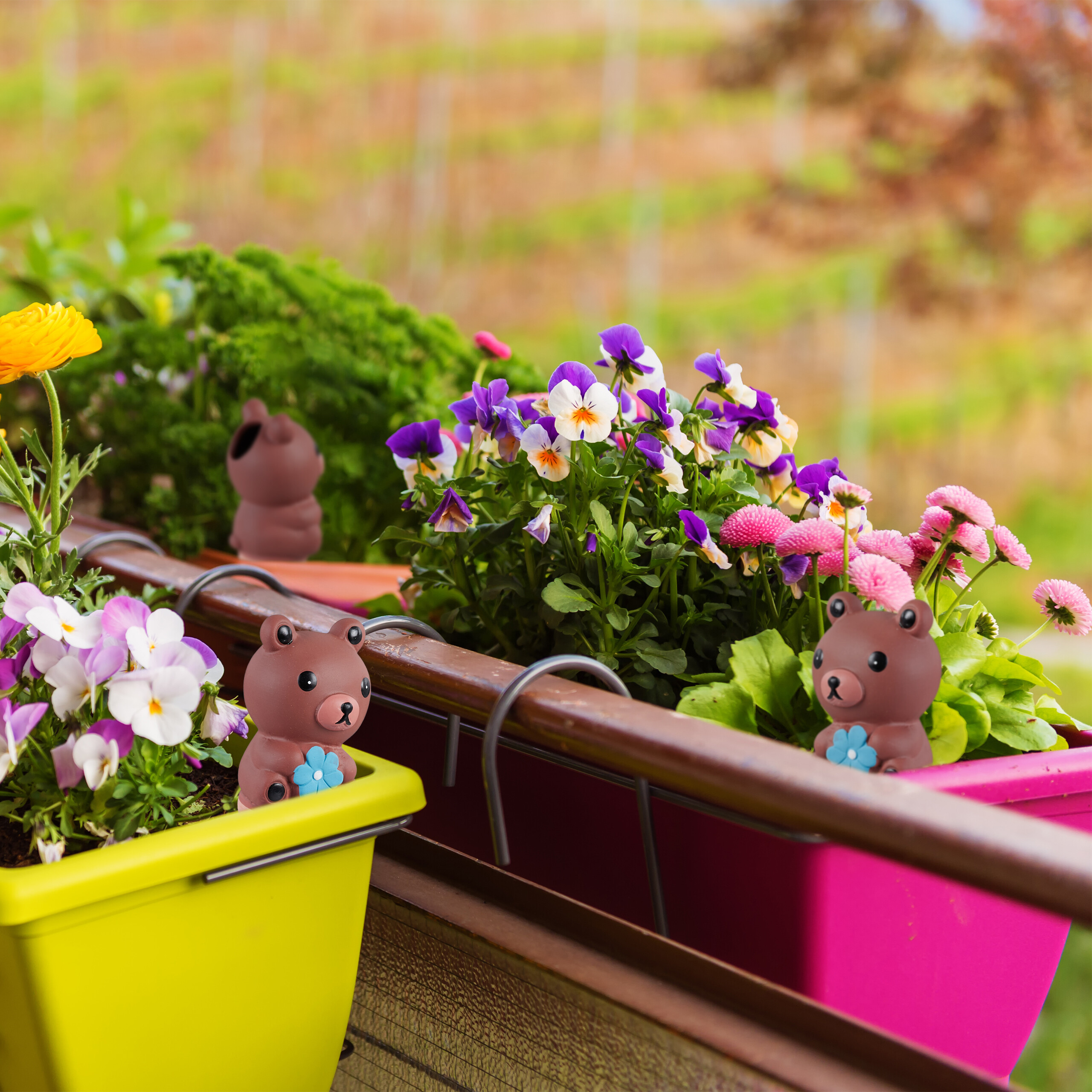 Bunte Blumen und kleine braune Bärenfiguren in Balkonkästen vor verschwommenem Hintergrund.