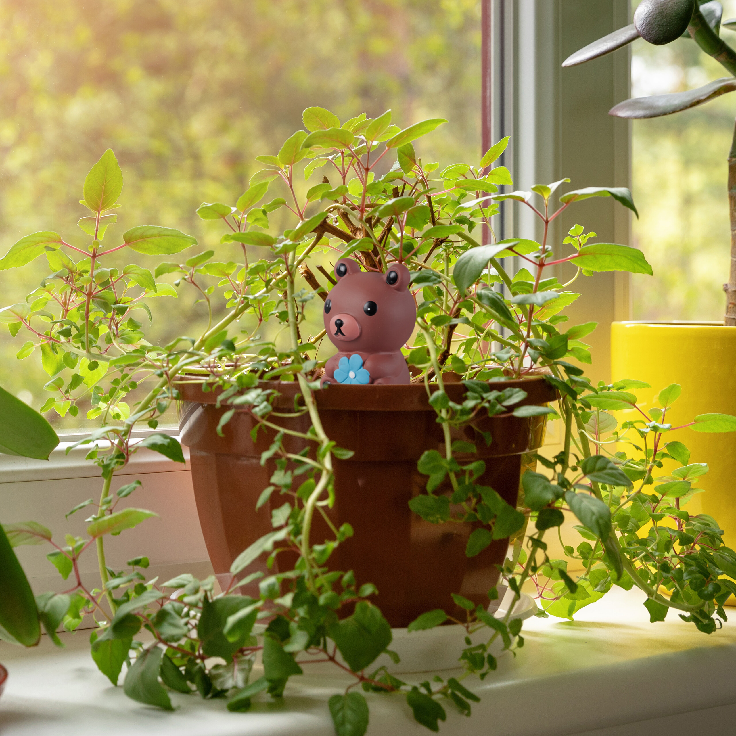 Ein kleiner brauner Bär sitzt in einem braunen Blumentopf voller grüner Pflanzen auf einer Fensterbank.