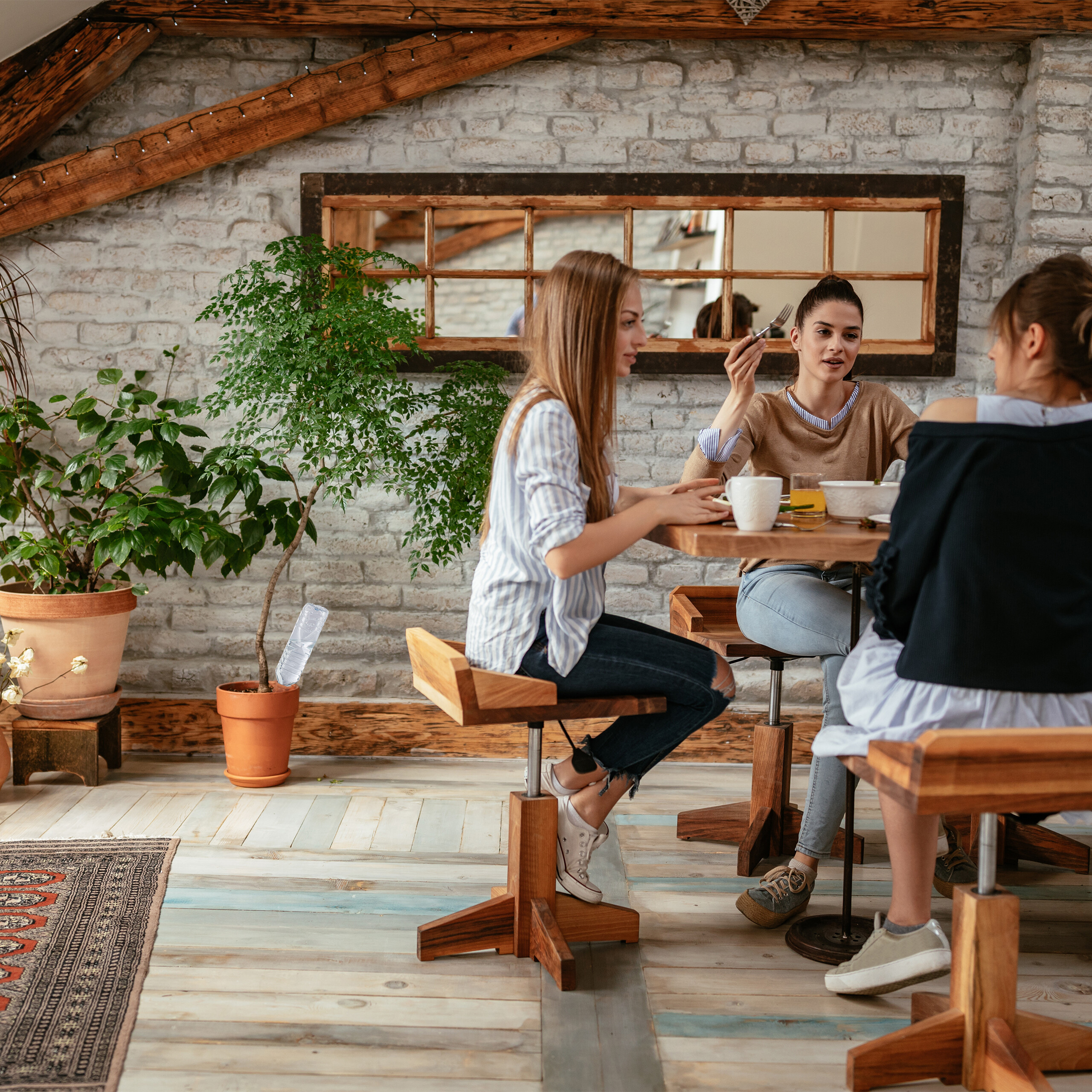 Drei Frauen sitzen an einem Holztisch im Loft mit Ziegelwand, Pflanzen und Spiegel.