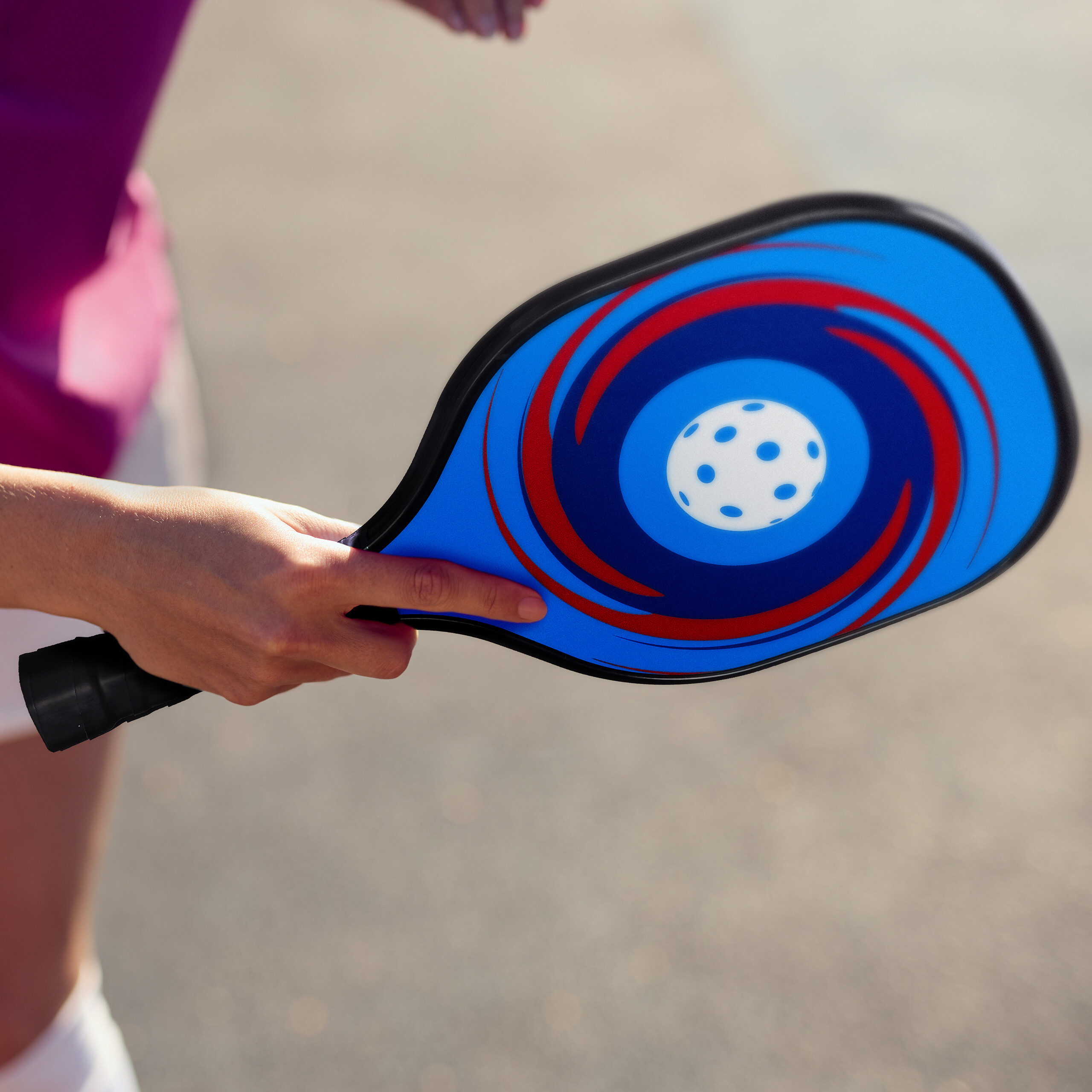 Eine Person hält einen blauen Pickleball-Schläger mit rotem Wirbeldesign und weißem Ball auf einem Außenplatz.