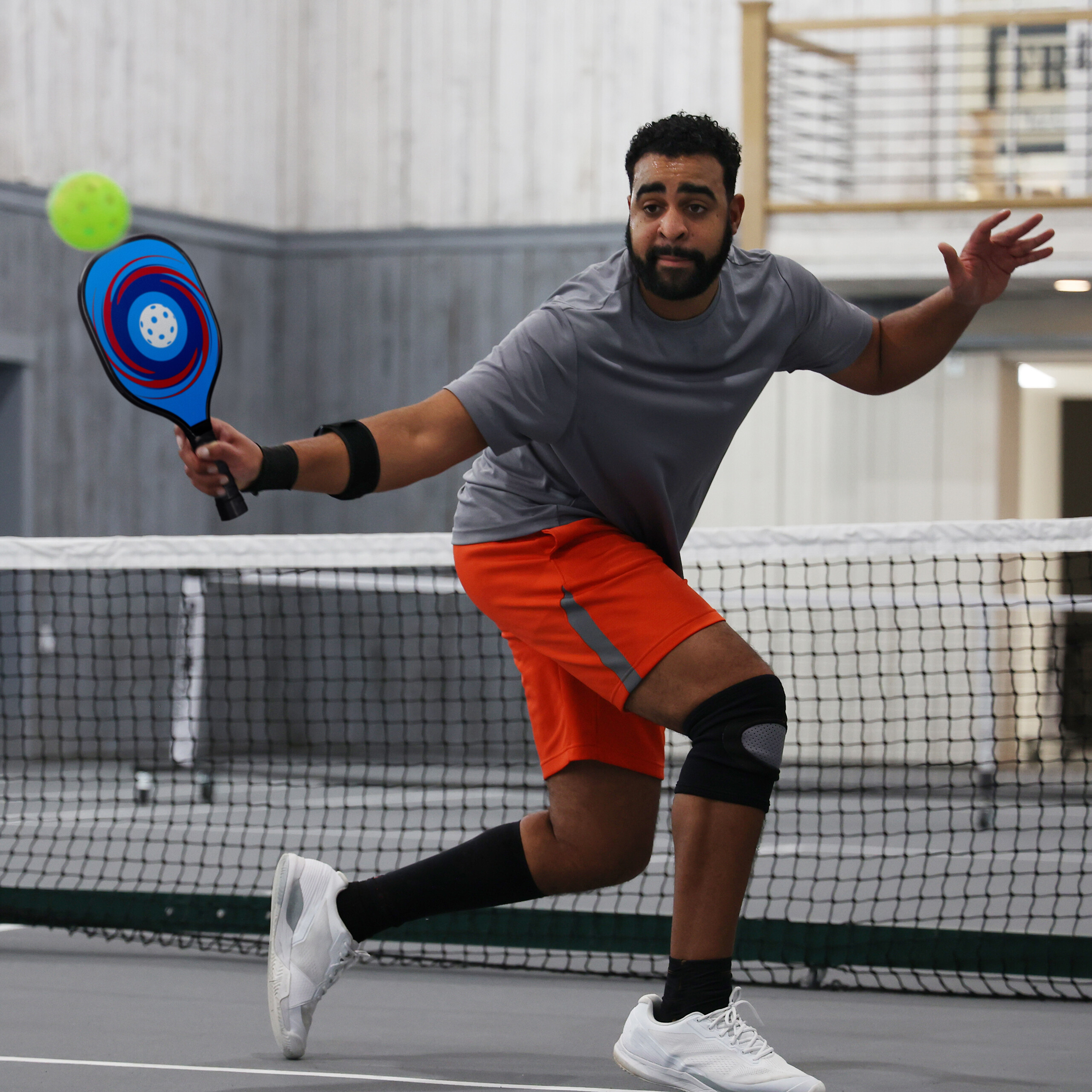Ein Mann in grauem Shirt und orangefarbenen Shorts spielt Pickleball und schlägt einen gelben Ball.