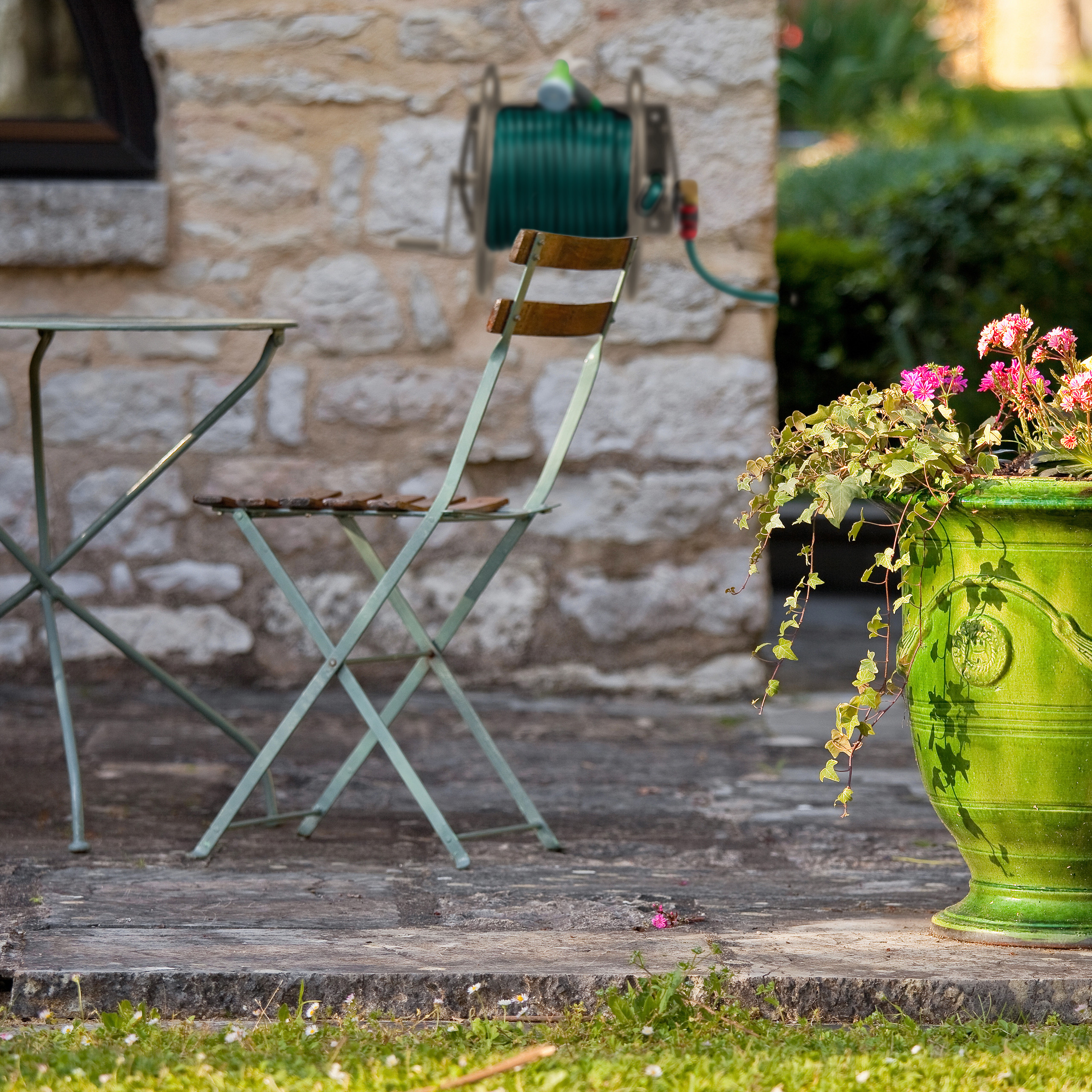 Ein grüner Topf mit Blumen auf einer Terrasse, im Hintergrund ein Tisch, Stühle und eine Schlauchtrommel an einer Steinwand.