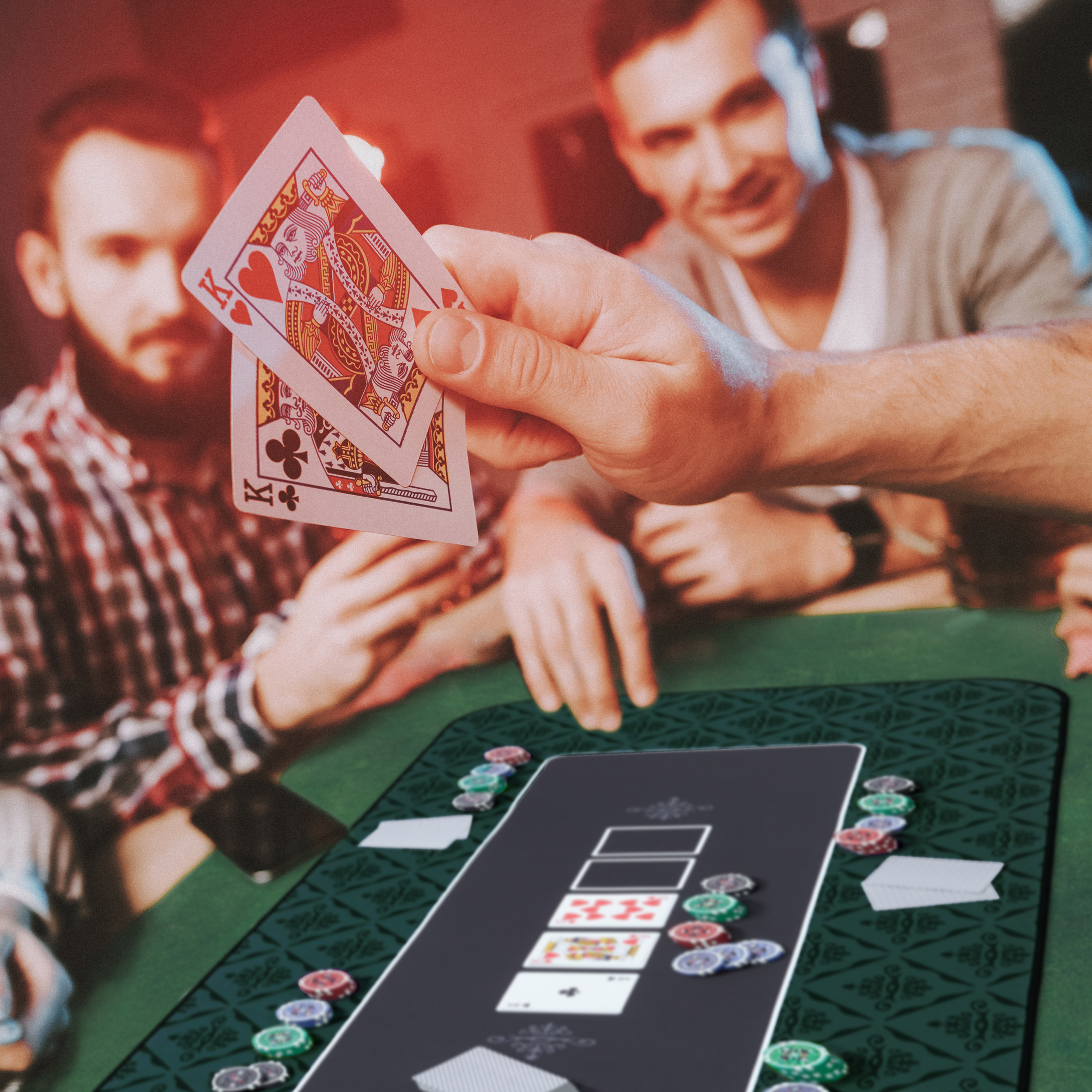 Ein Mann hält zwei Königskarten über einen Pokertisch, während im Hintergrund zwei andere Männer spielen.