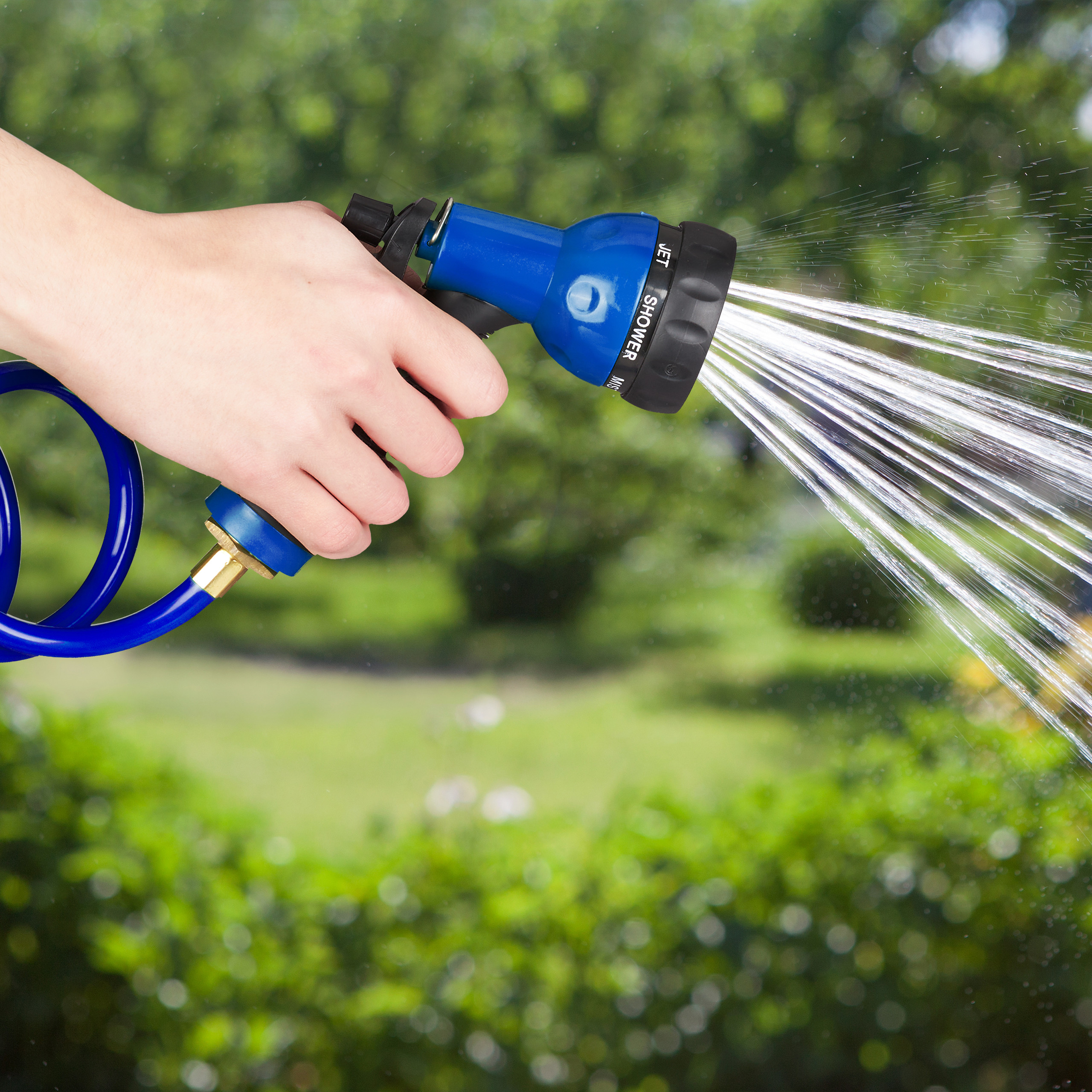 Eine Hand hält eine blaue Garten-Spritzpistole, aus der Wasser strömt, mit unscharfem Gartenhintergrund.