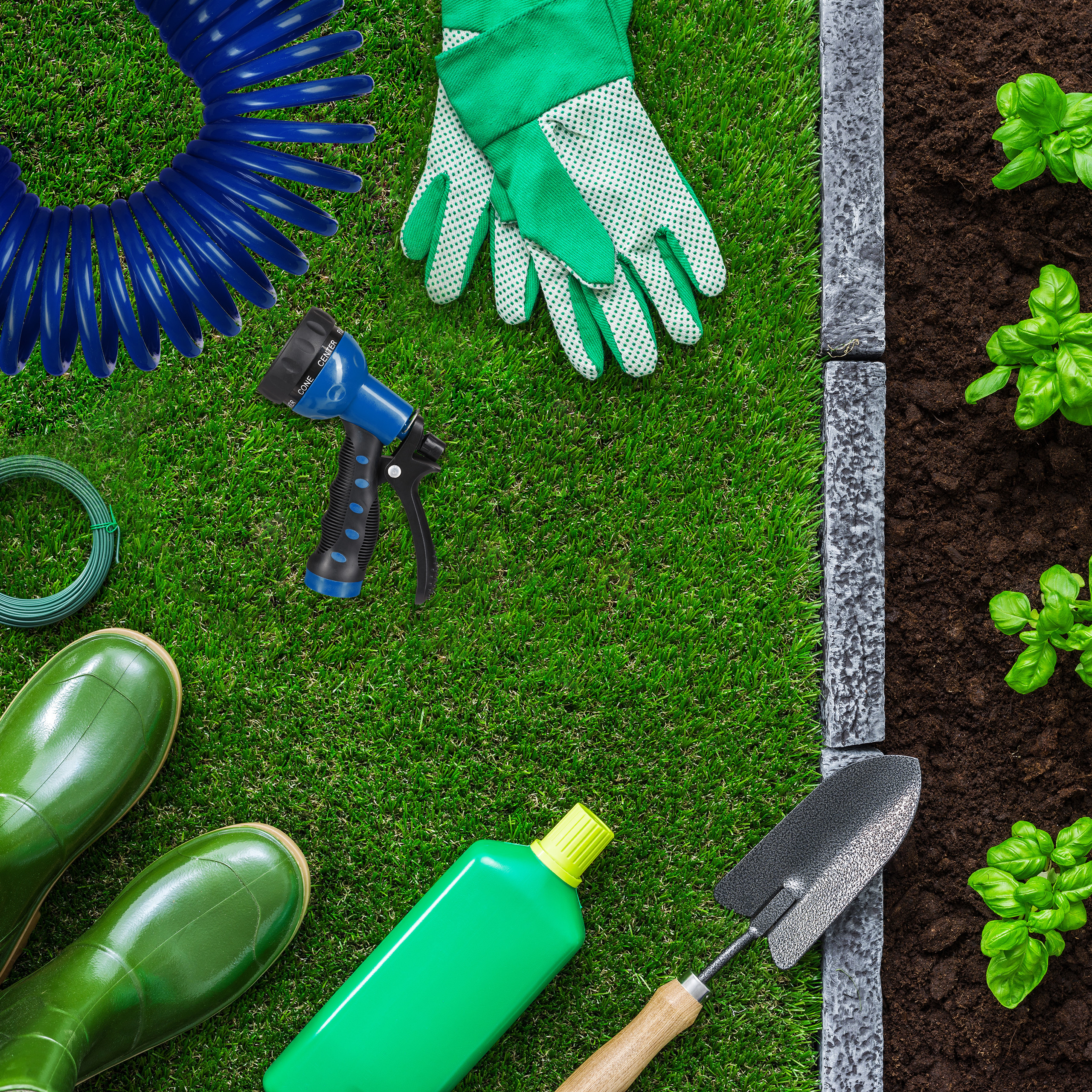 Gartengeräte wie Schlauch, Handschuhe, Stiefel und Kelle auf Gras neben einem Beet.