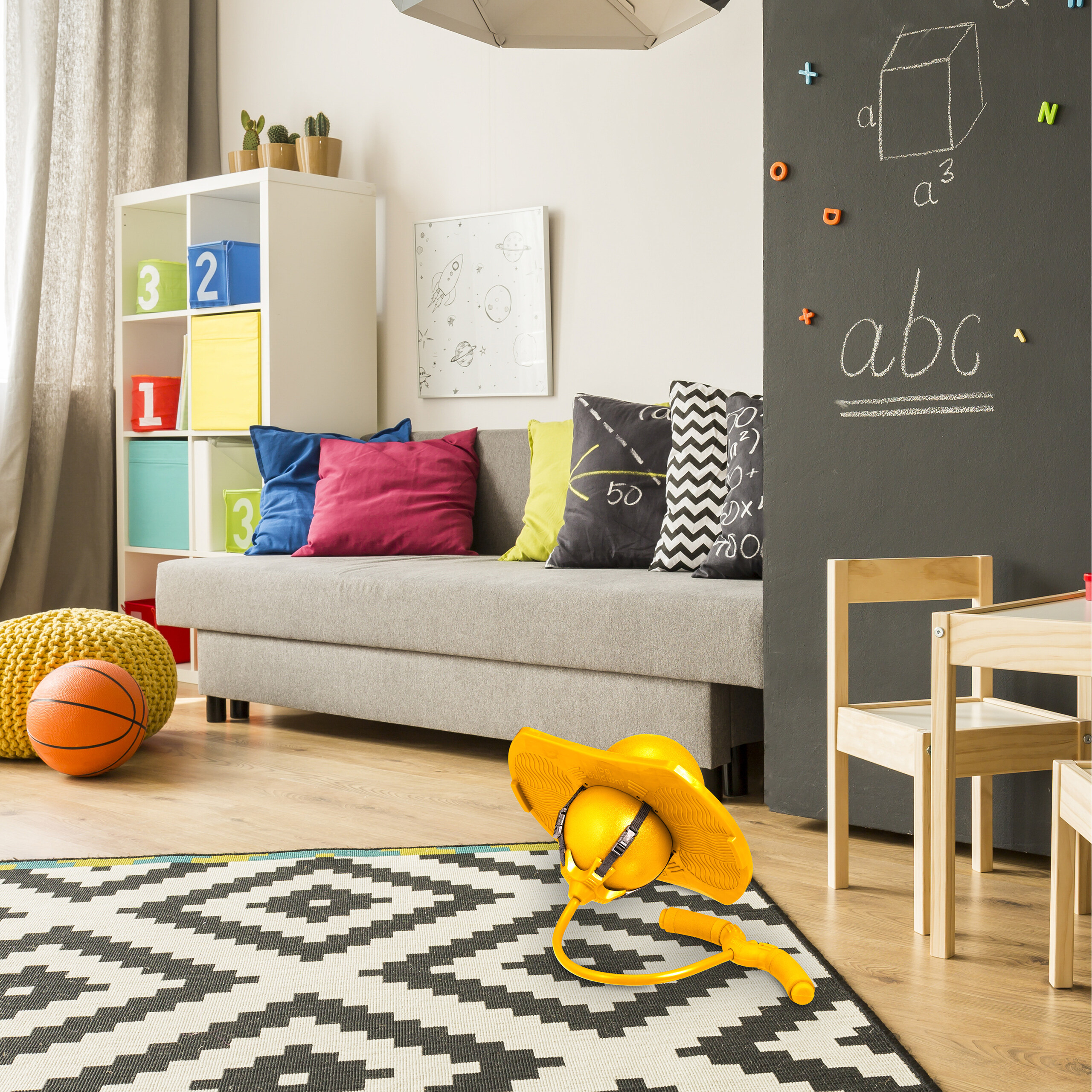 Buntes Kinderzimmer mit grauem Sofa, Teppich, Pogo-Stick und großer Wandtafel.