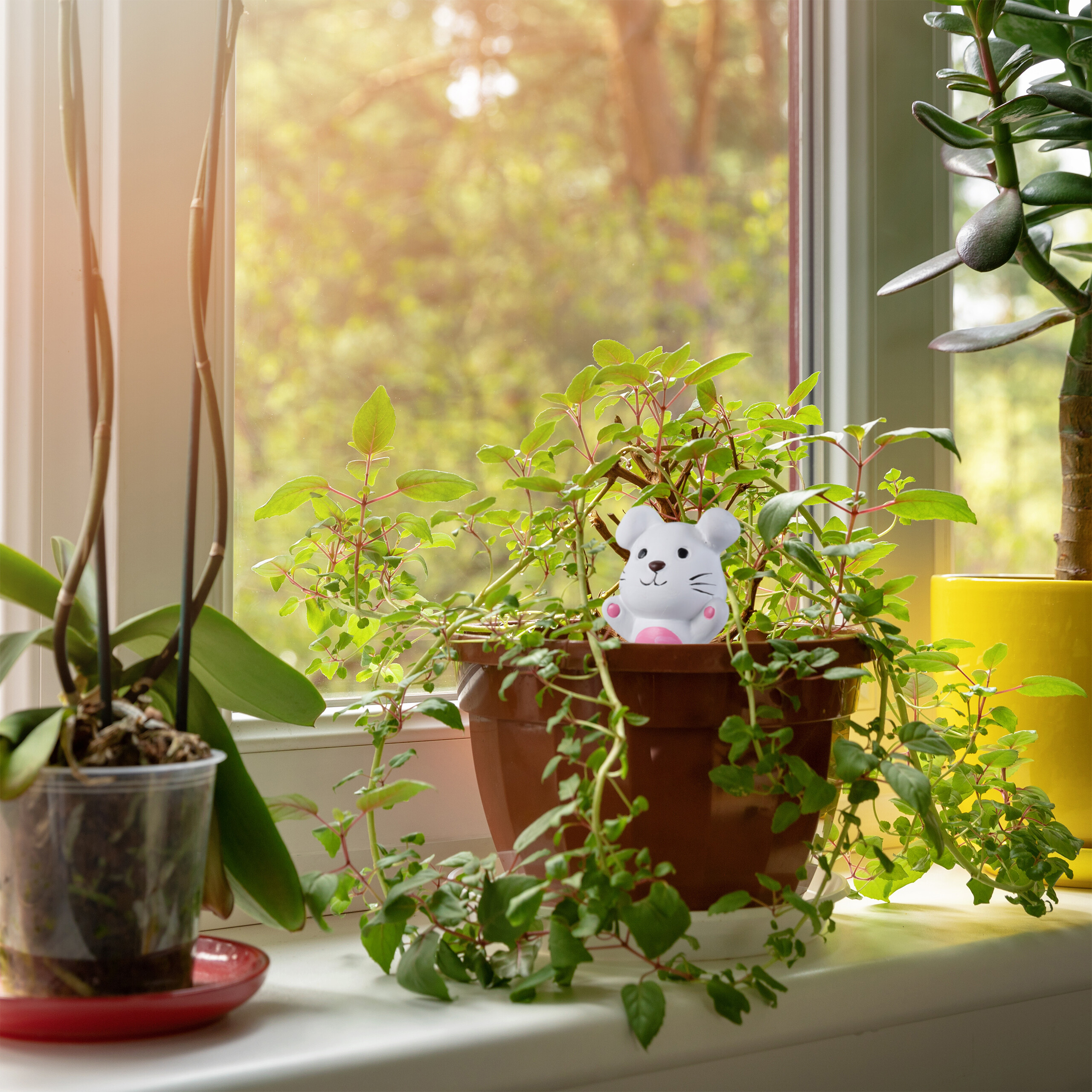 Eine kleine weiße Mausfigur sitzt in einem braunen Blumentopf auf einer sonnigen Fenstersbank zwischen grünen Pflanzen.