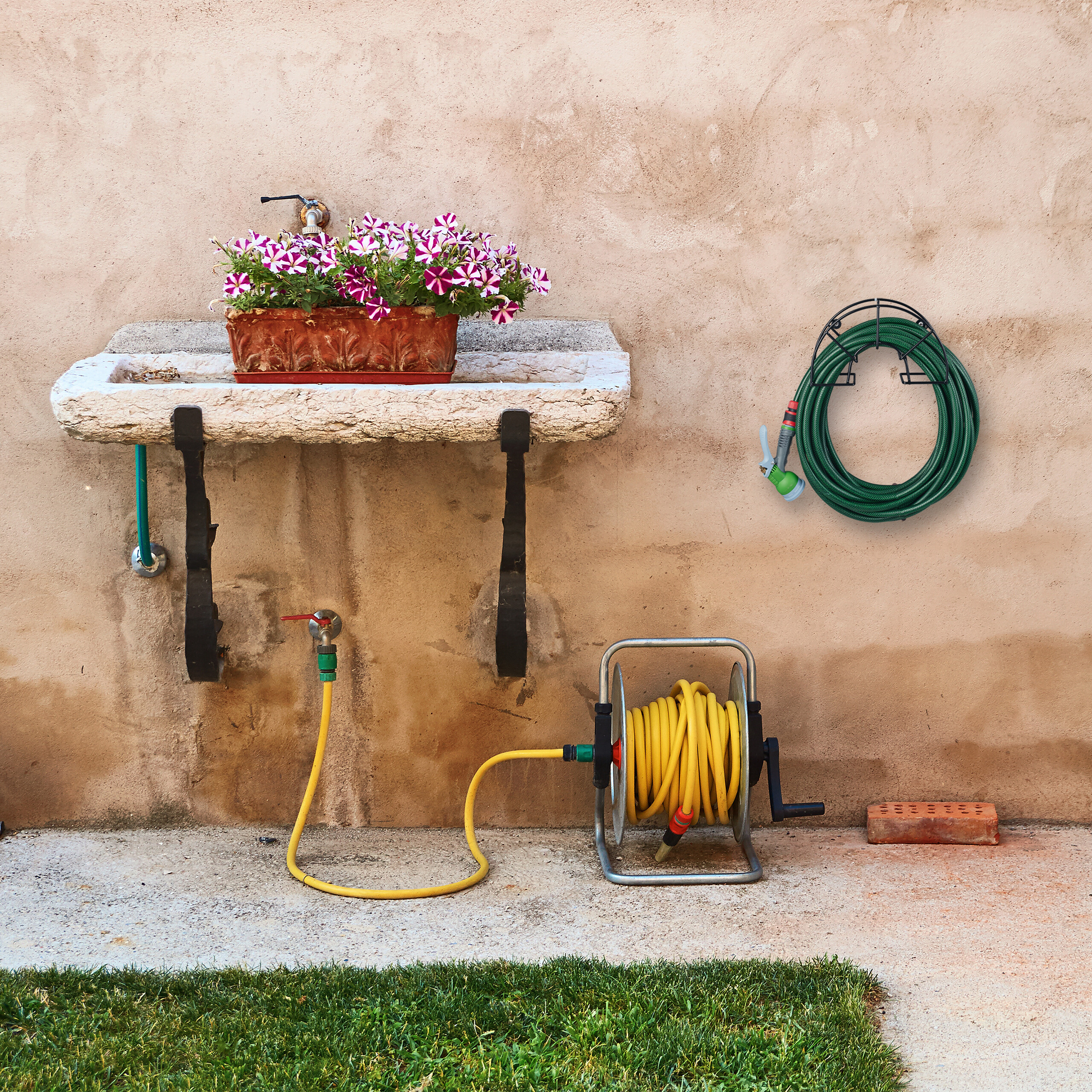 Ein Steintrog mit Blumen, ein Wasserschlauch auf einer Rolle und ein Wandhalter an einer beigefarbenen Wand.