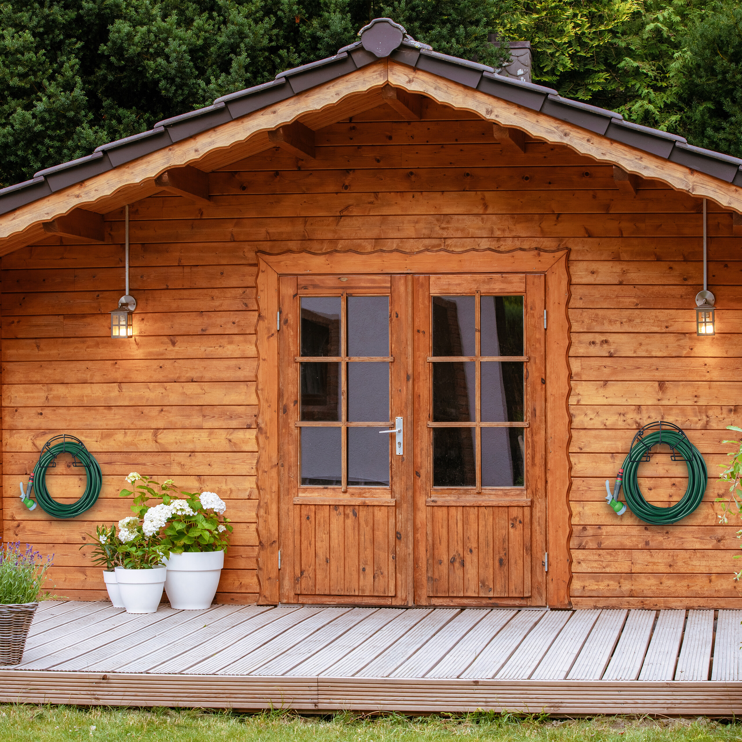 Eine Gartenlaube aus Holz mit Doppeltüren, Veranda, Blumentöpfen und zwei grünen Schläuchen an den Wänden.