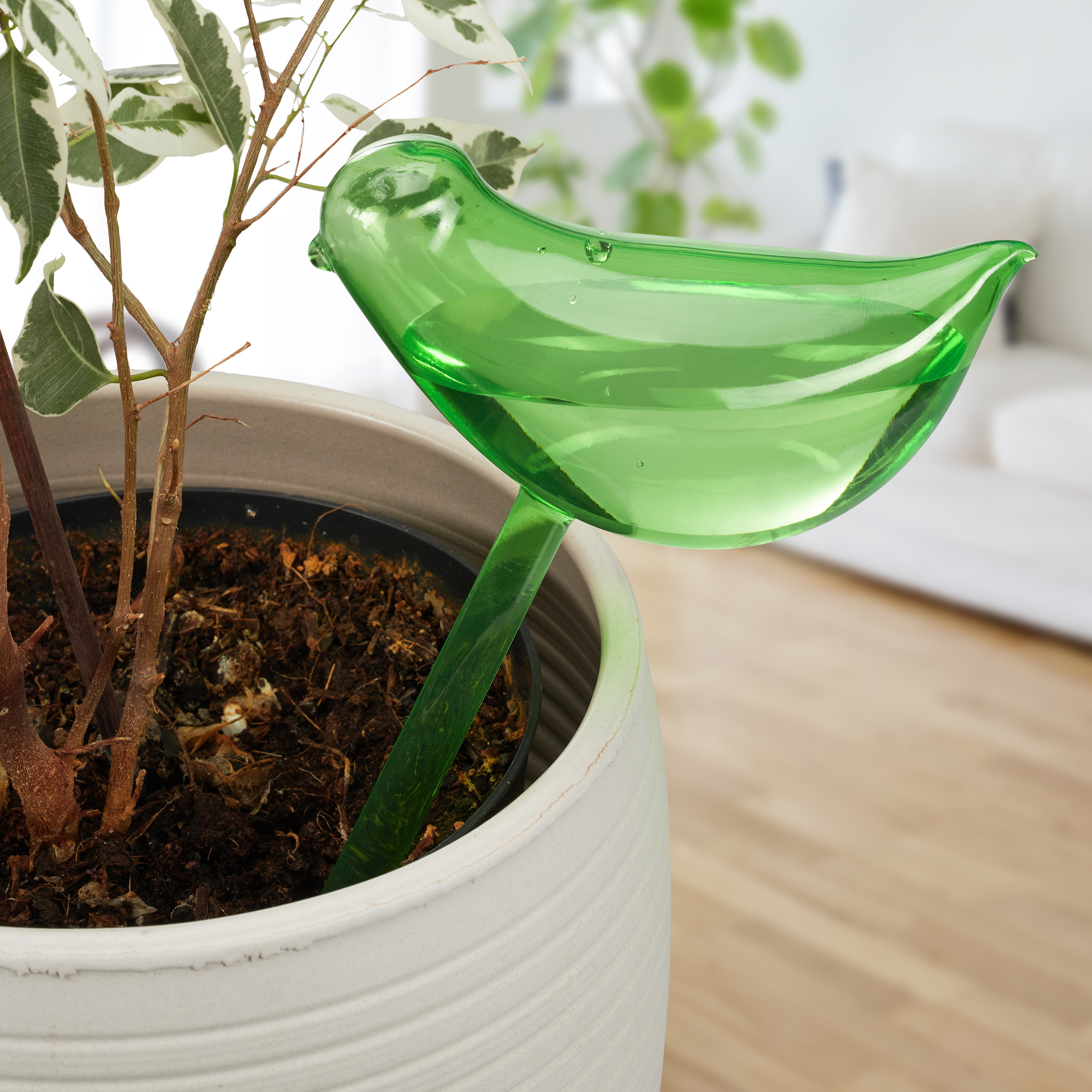 Ein grüner Gießkugel-Vogel aus Glas steckt in der Erde eines Topfhekens.
