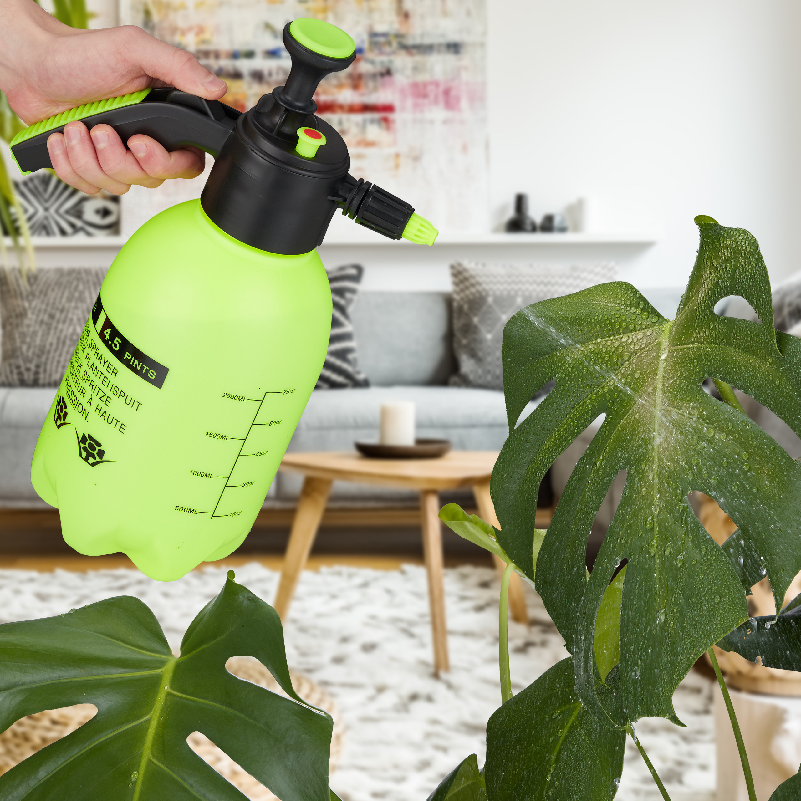 Eine Hand hält eine neongrüne Pflanzensprühflasche und besprüht eine Monstera-Pflanze in einem Wohnzimmer.