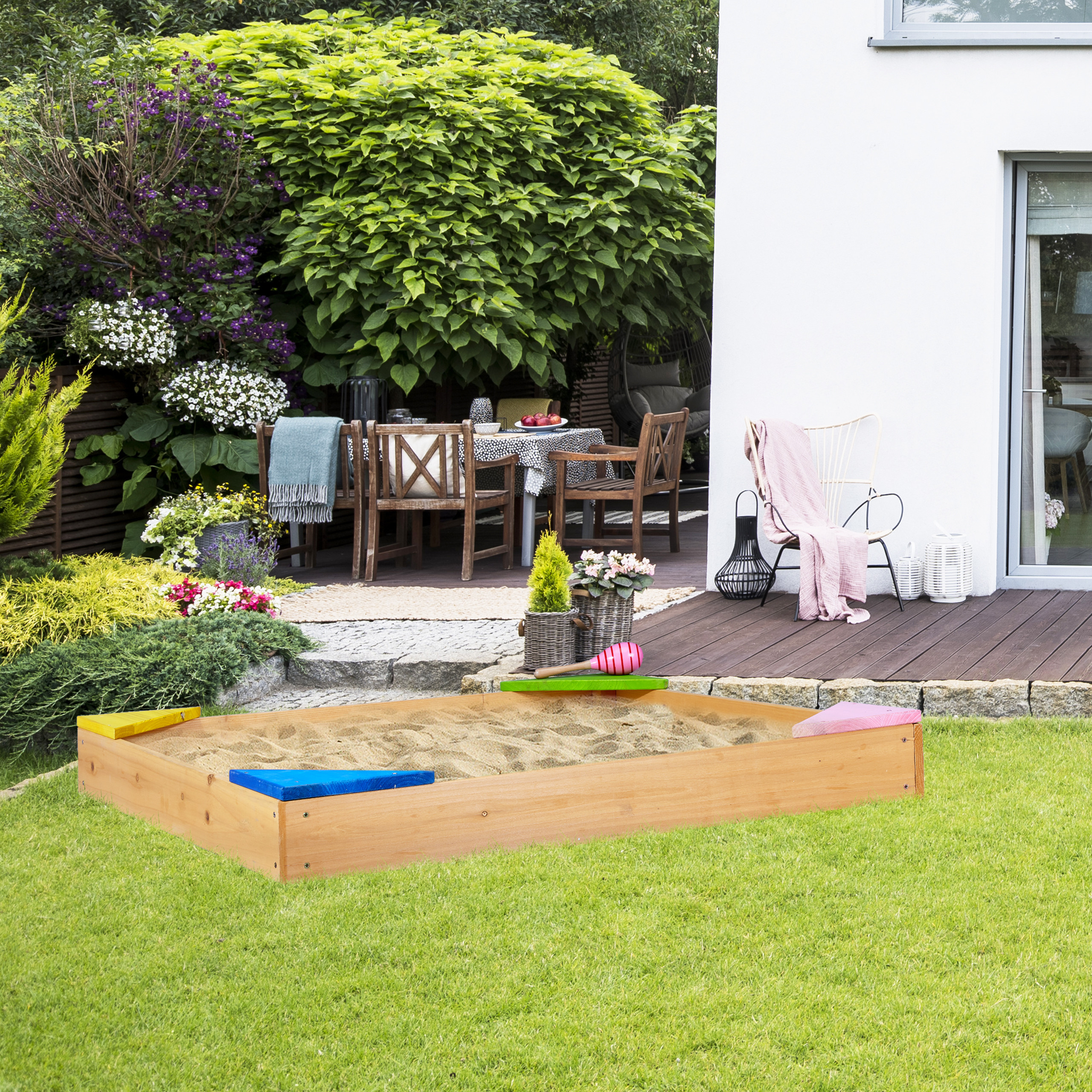 Ein hölzerner Sandkasten mit bunten Ecksitzen auf einem grünen Rasen im Garten mit Terrasse und üppiger Bepflanzung.