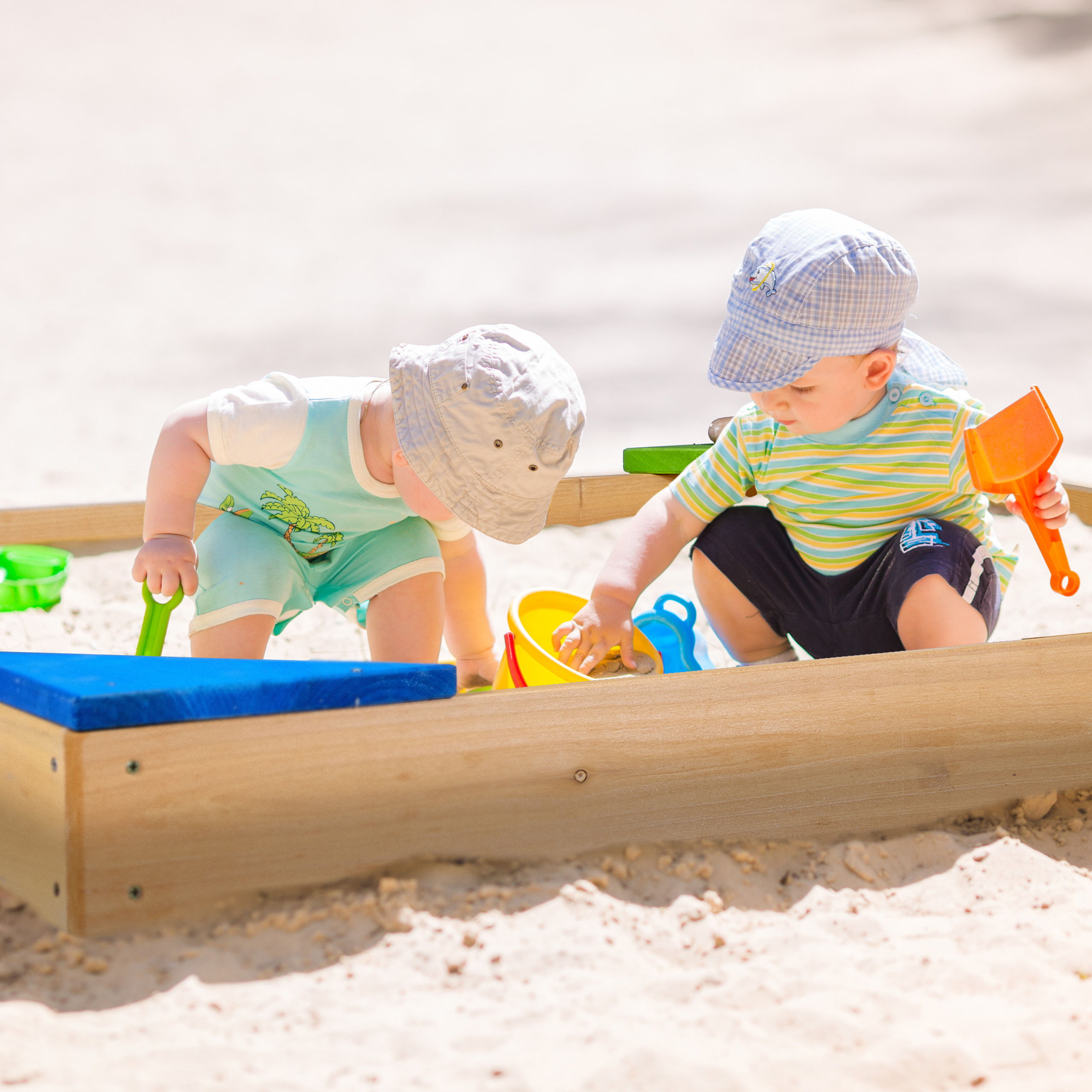 Zwei Kleinkinder mit M ützen spielen an einem sonnigen Tag mit Spielzeug und Sand in einem Holzkasten.