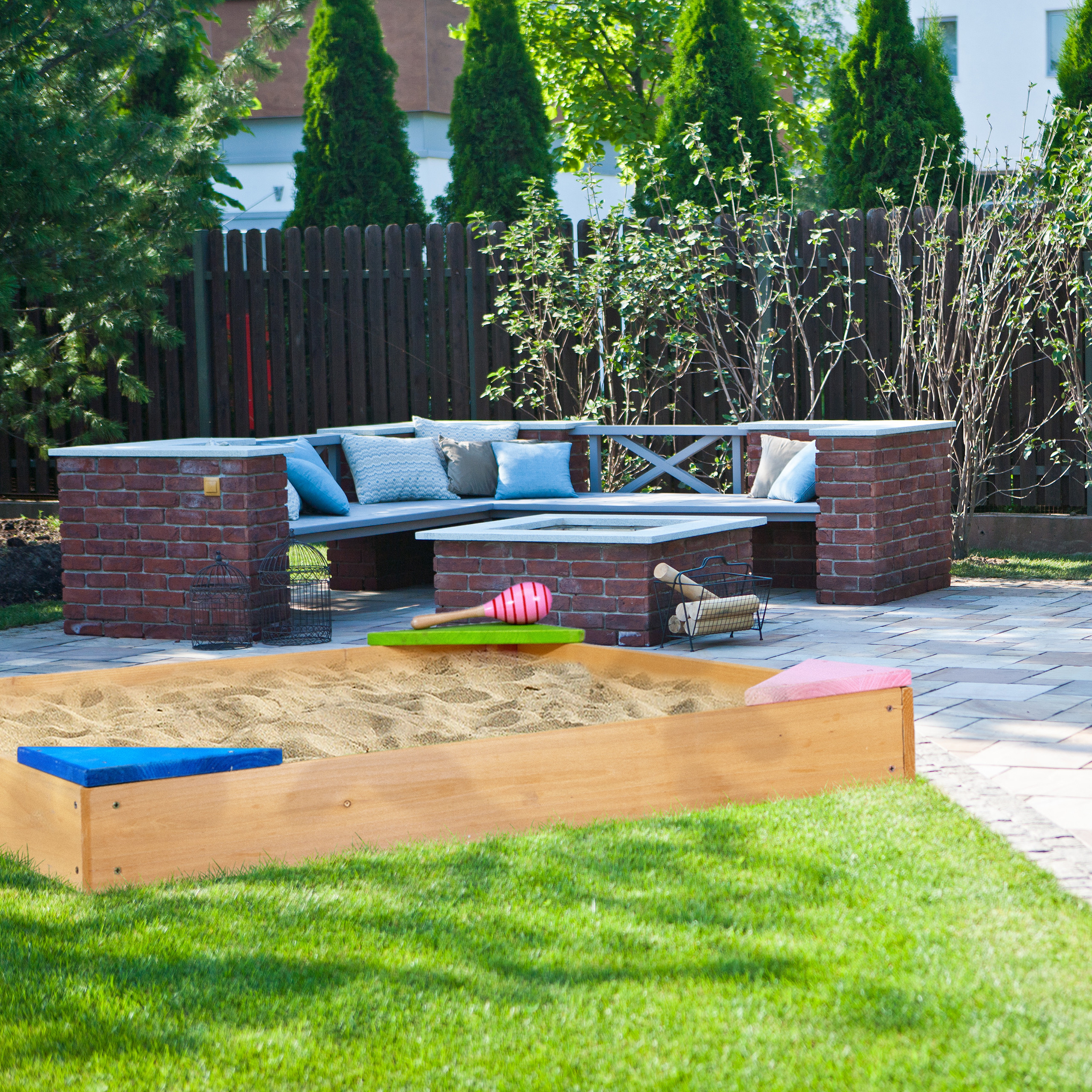 Ein Holzsandkasten mit Sand und Spielzeug steht auf Rasen in einem Garten mit Backsteinsitzbereich und dunklem Zaun.