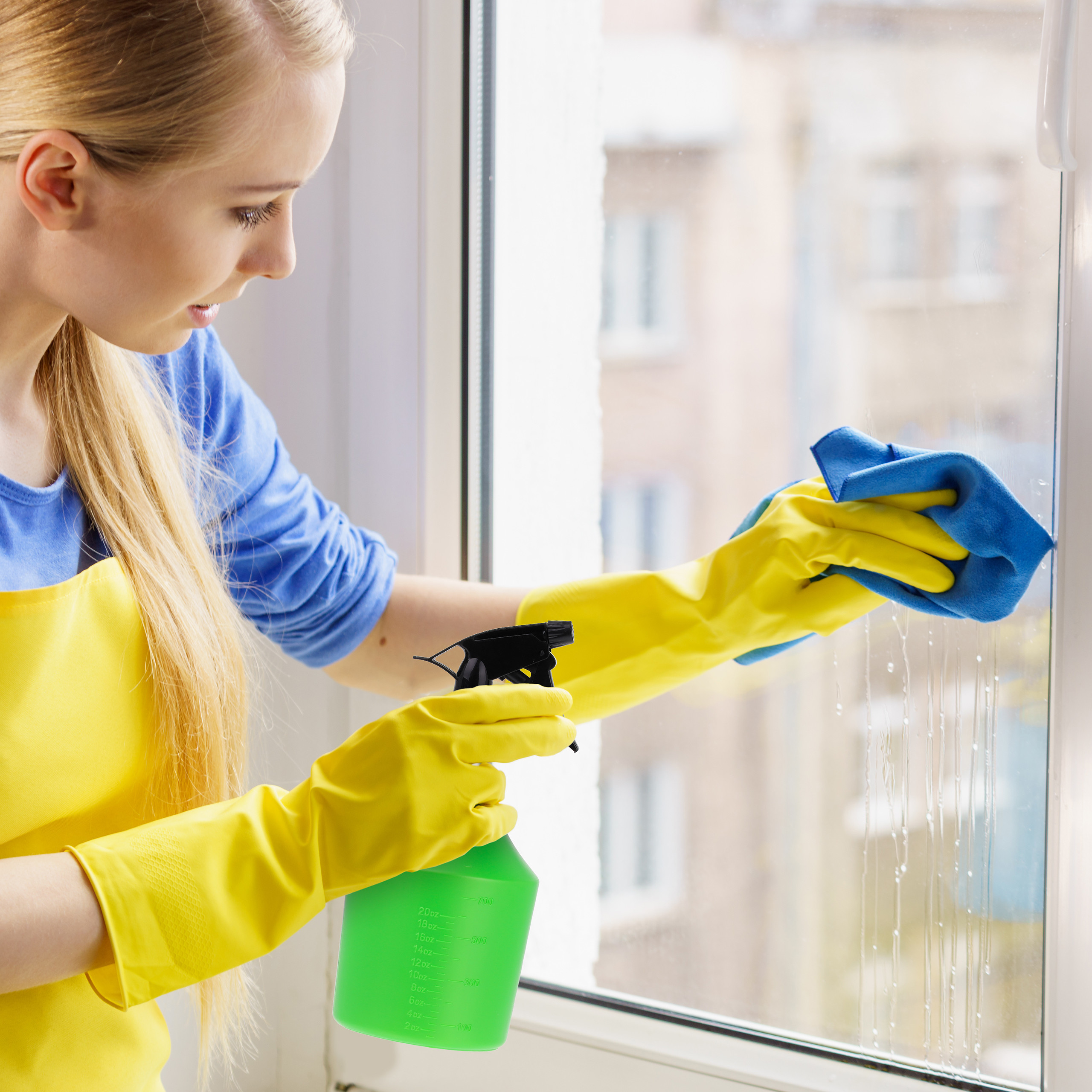 Blonde Frau mit gelben Handschuhen putzt ein Fenster mit Reiniger und blauem Tuch.