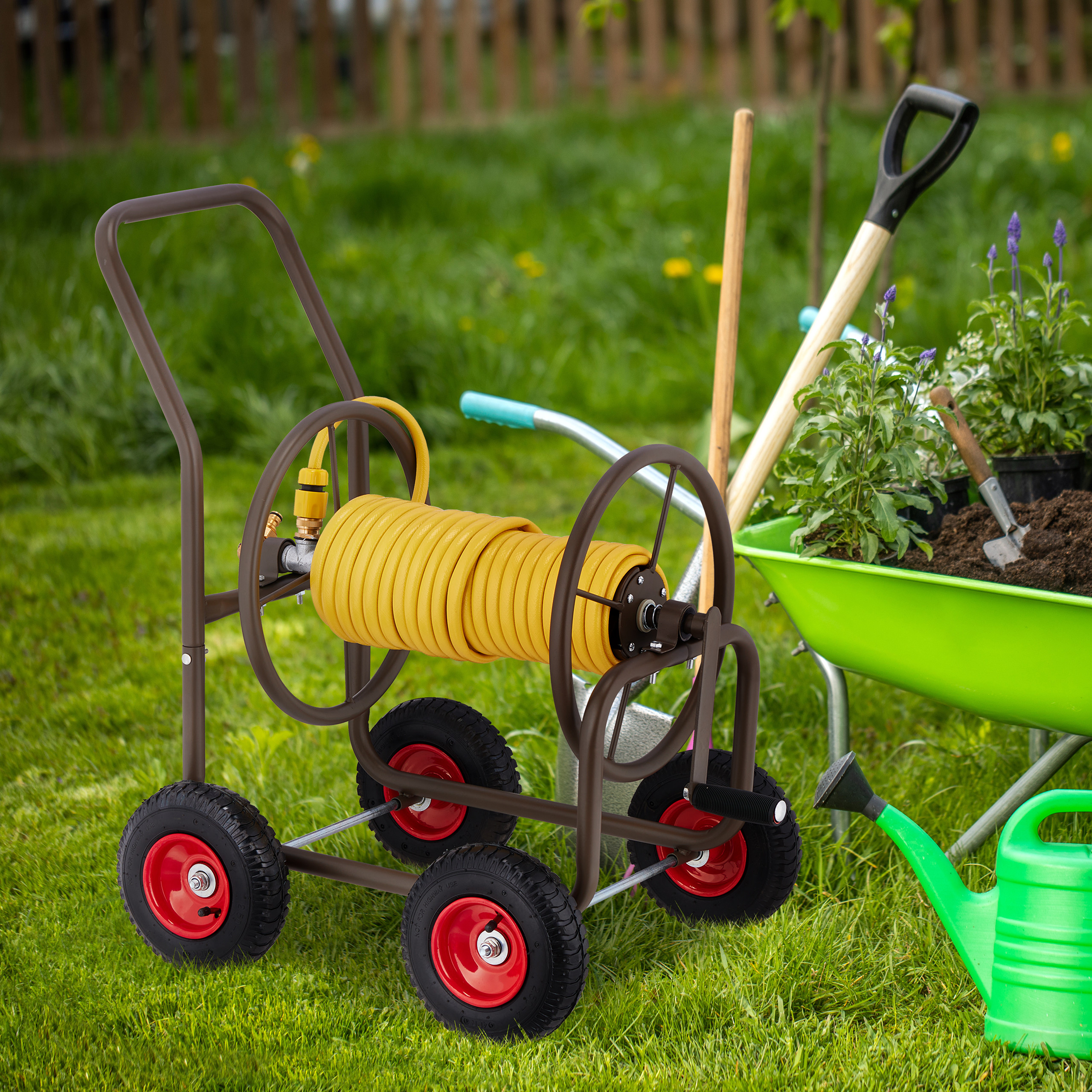 Ein Gartenschlauchwagen mit gelbem Schlauch auf Rasen, neben einer grünen Schubkarre und Werkzeugen.