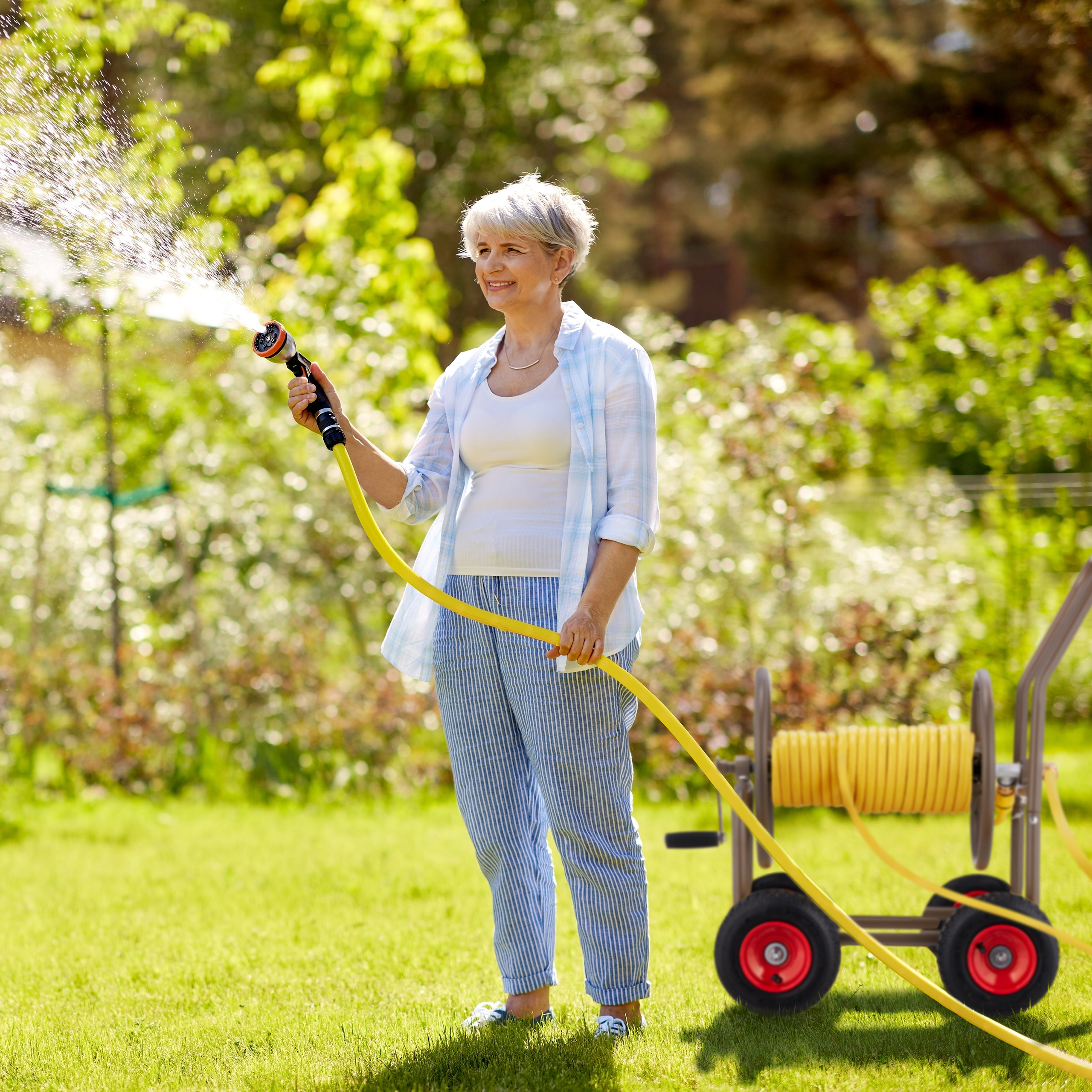 Eine ältere Frau gießt den Rasen mit einem gelben Gartenschlauch in einem sonnigen Garten.