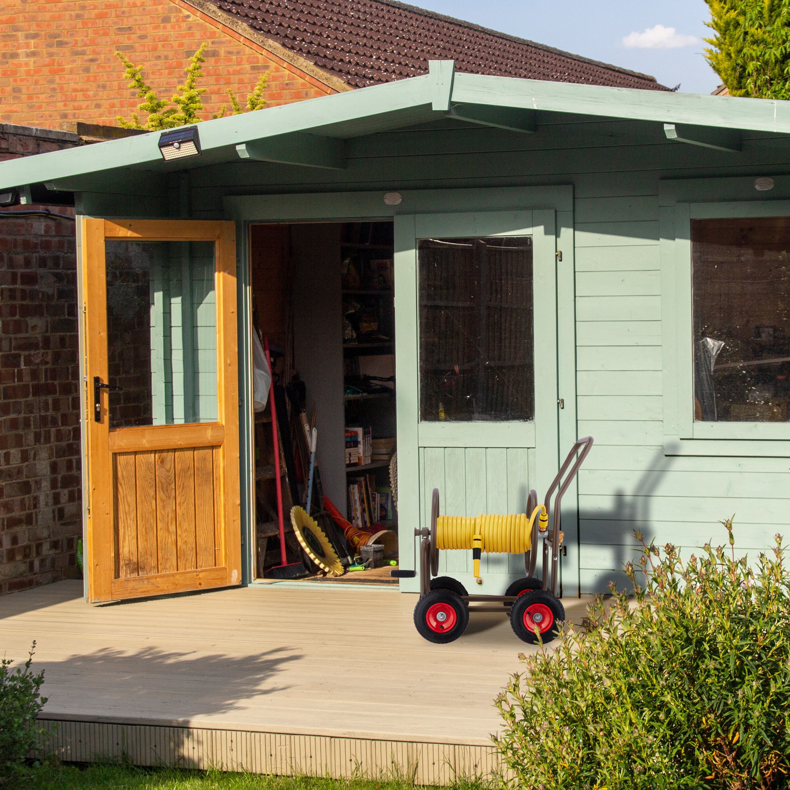 Ein hellgrünes Gartenhaus mit offener Holztür, Regalen im Inneren und einem gelben Schlauchwagen auf dem Holzdeck.