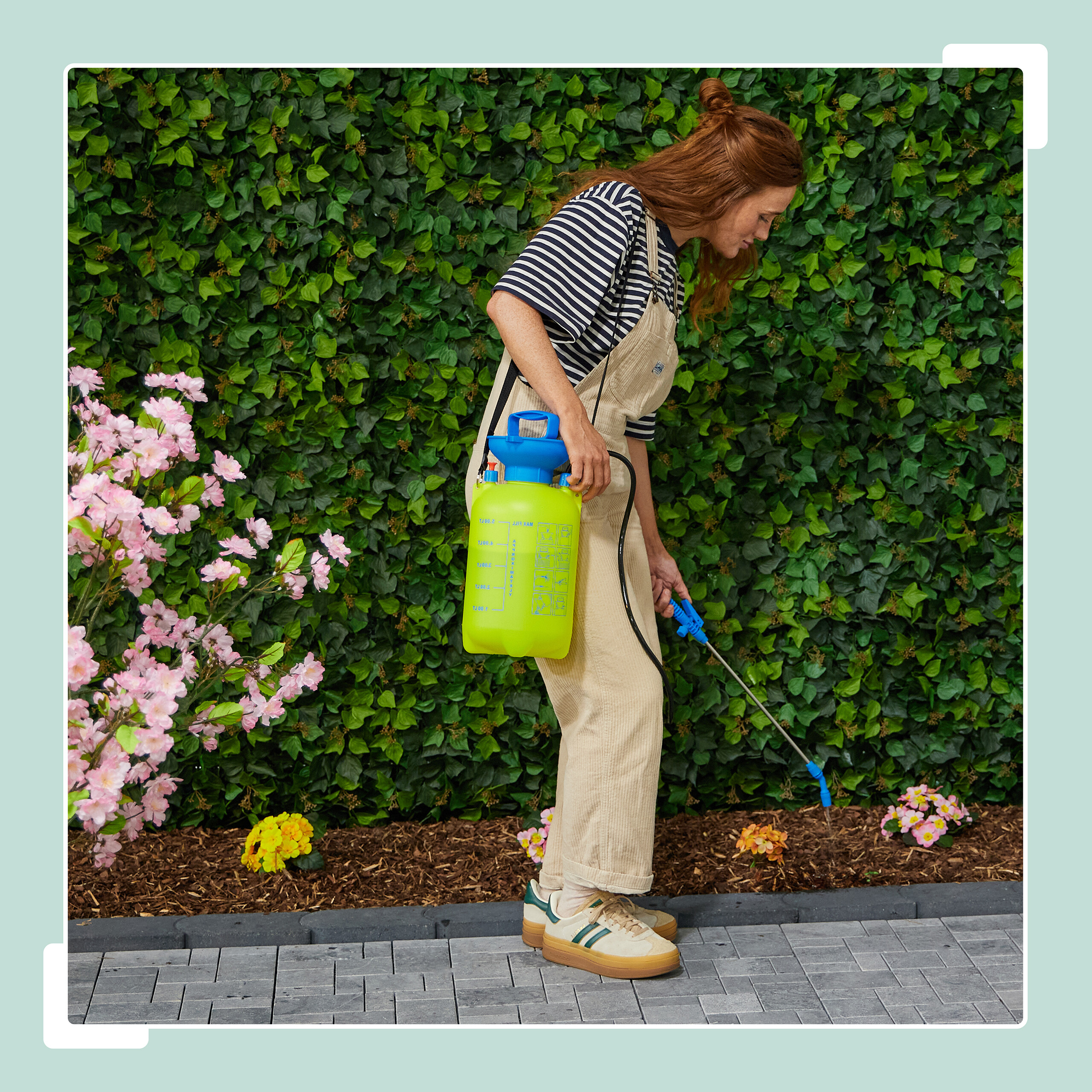 Eine Frau in Latzhose sprüht mit einem grünen Drucksprüher Pflanzen vor einer grünen Hecke.