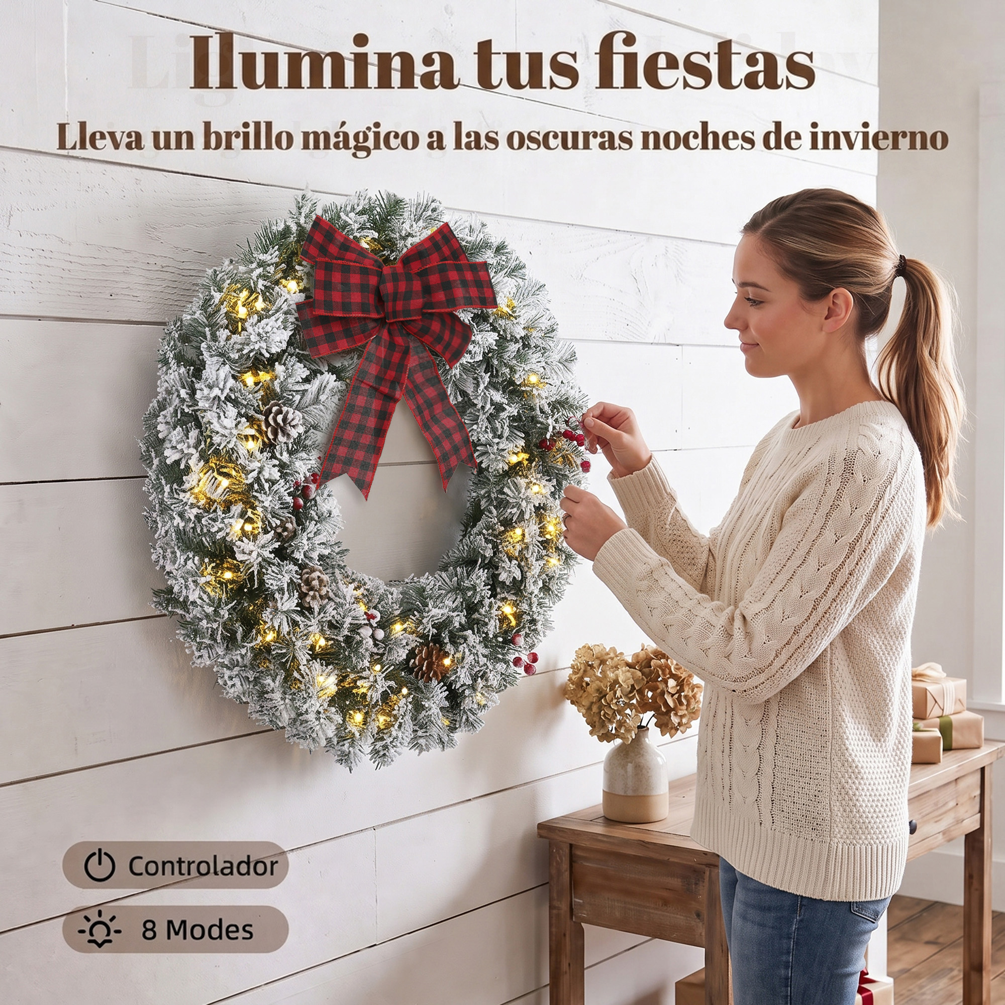 Una mujer con suéter blanco cuelga una corona navideña iluminada con un lazo rojo en una pared.