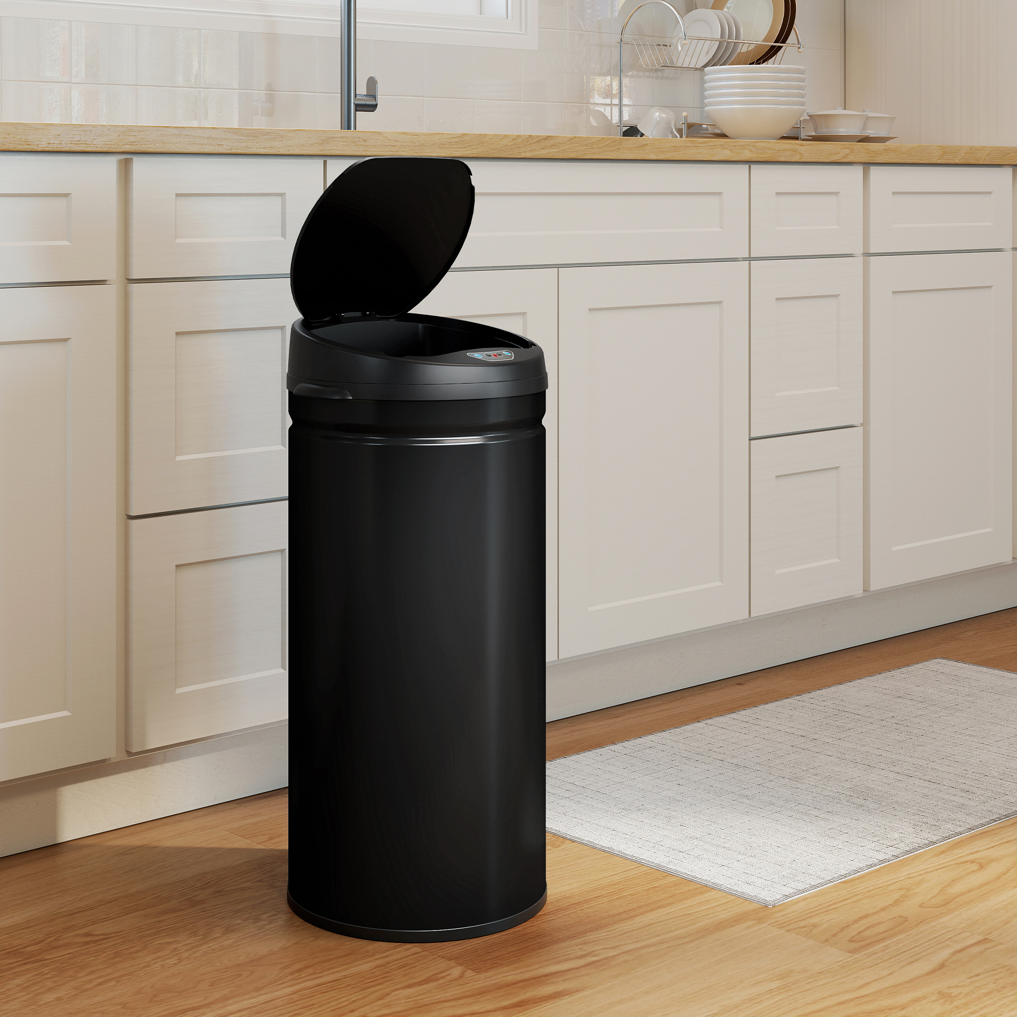 Un cubo de basura negro con la tapa abierta se encuentra en un suelo de madera frente a armarios de cocina blancos.