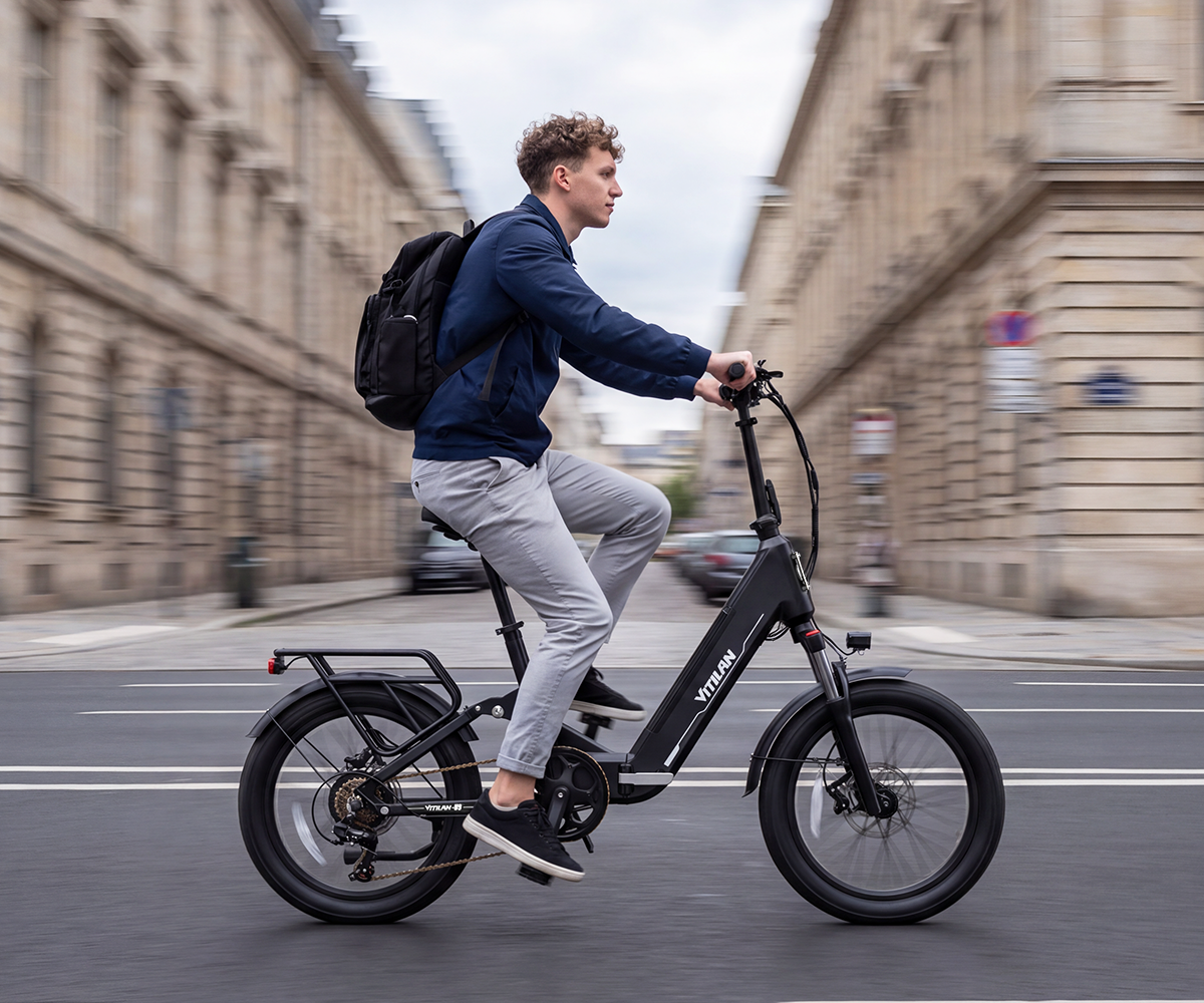 Un hombre con mochila monta en bicicleta eléctrica negra por una calle urbana entre edificios de piedra.
