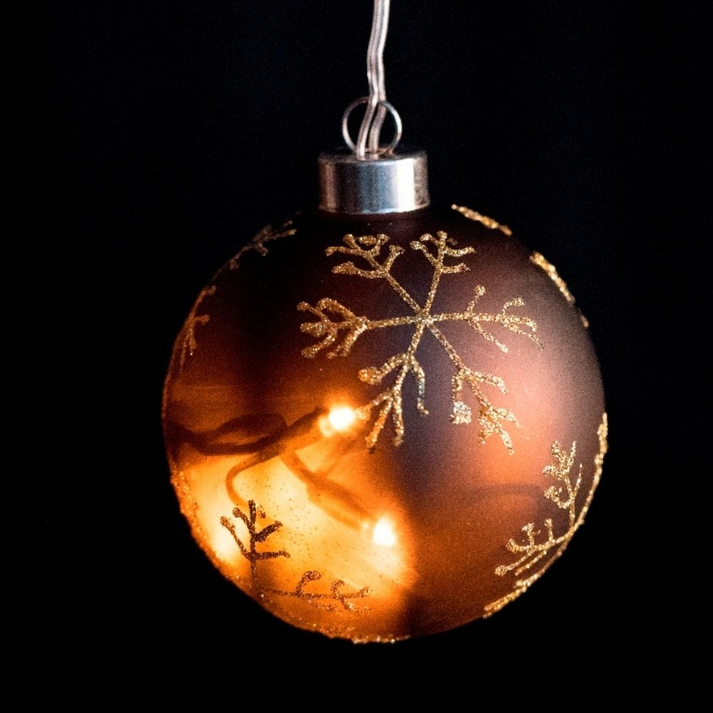 Un adorno navideño marrón con copos de oro y luces cálidas en el interior sobre fondo negro.