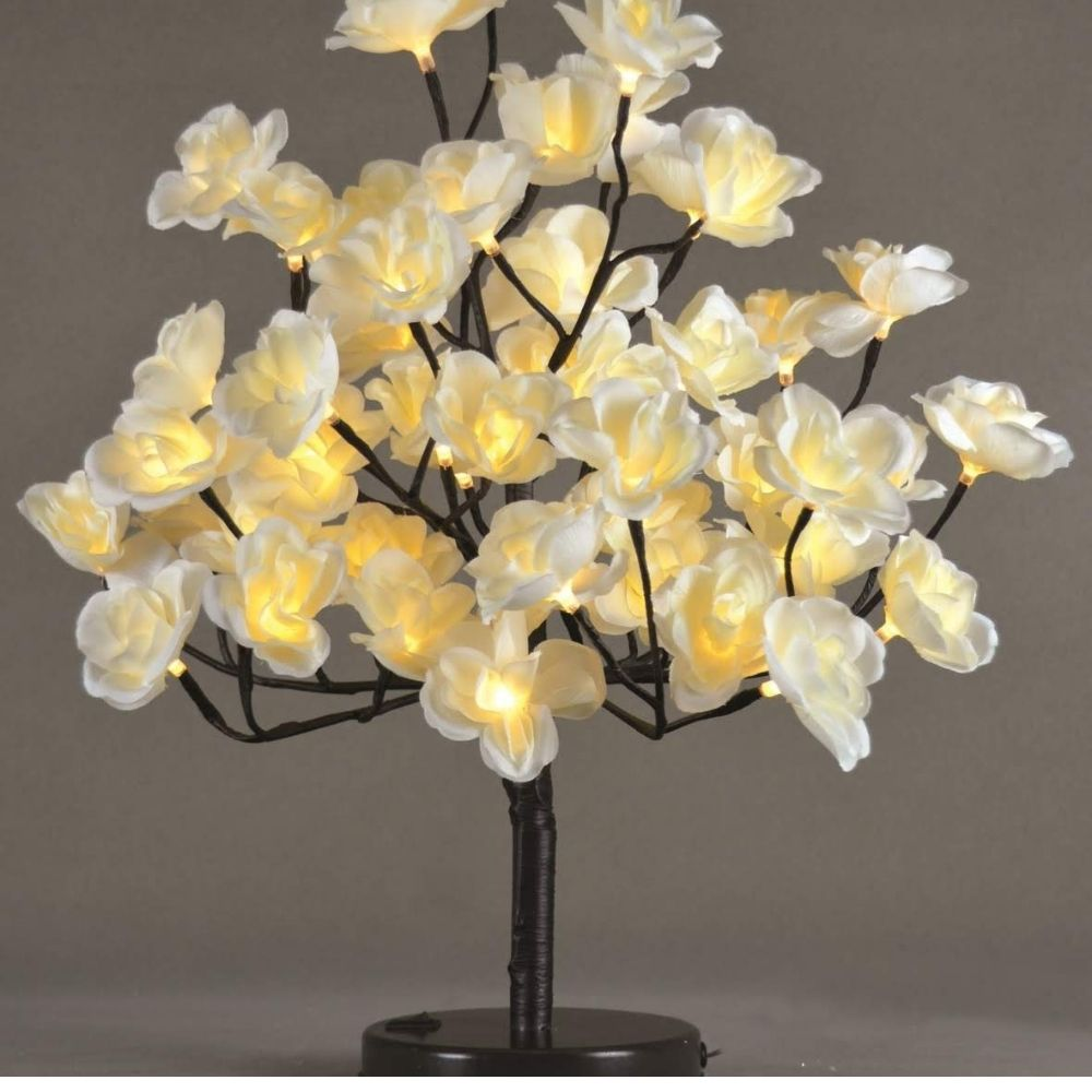 Una lámpara de mesa decorativa con luces blancas en forma de flor sobre ramas oscuras y un fondo gris liso.