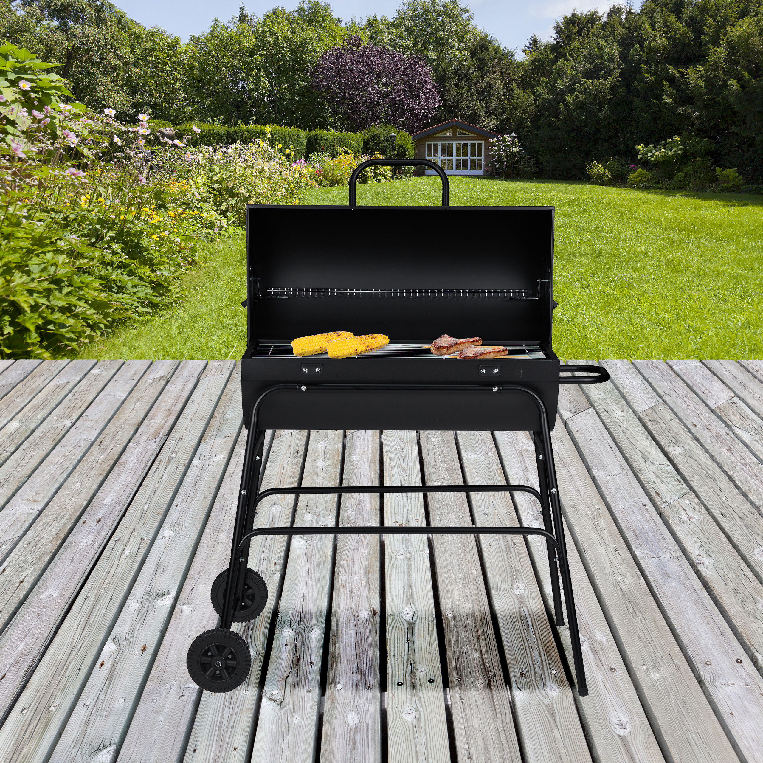 Ein schwarzer Holzkohlegrill auf einer Holzterrasse mit Mais und Fleisch auf dem Rost und Blick auf einen grünen Garten.